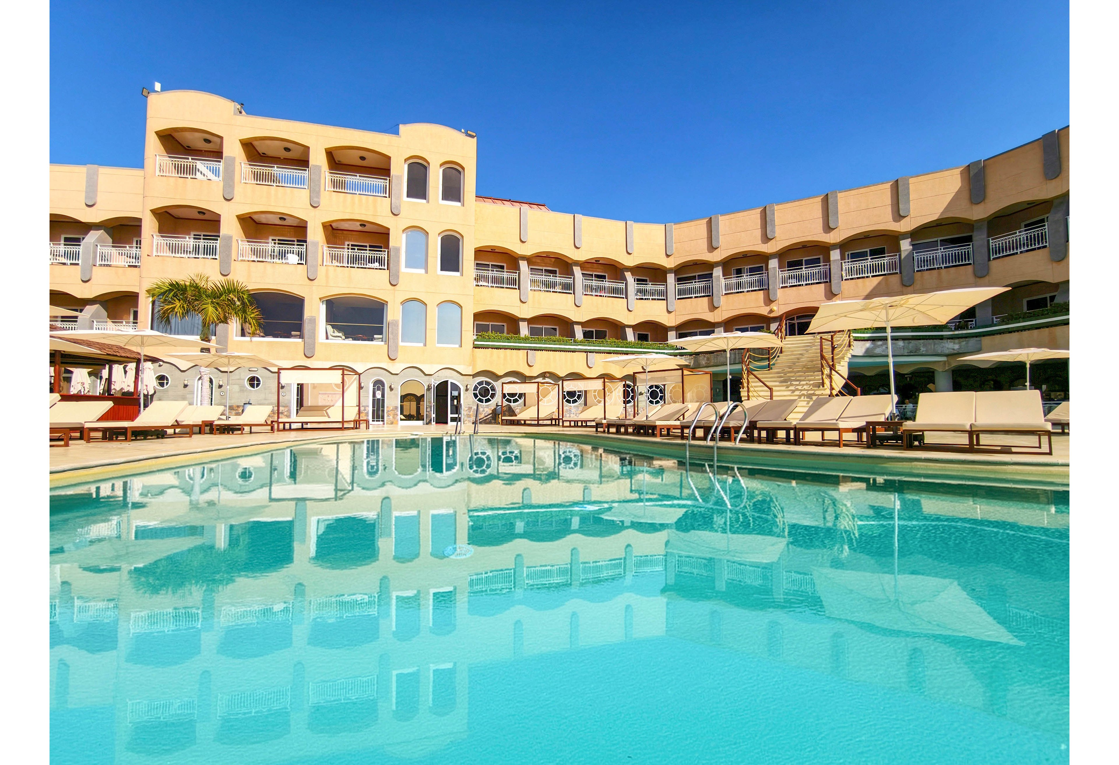 A large resort hotel curves around a bright blue outdoor swimming pool under a clear sky. Multiple balconies overlook the pool area, offering views of the water and sun loungers. White lounge chairs and umbrellas line the poolside deck, suggesting a relaxing vacation atmosphere. The calm water reflects the building’s facade, enhancing the sense of symmetry and tranquility.