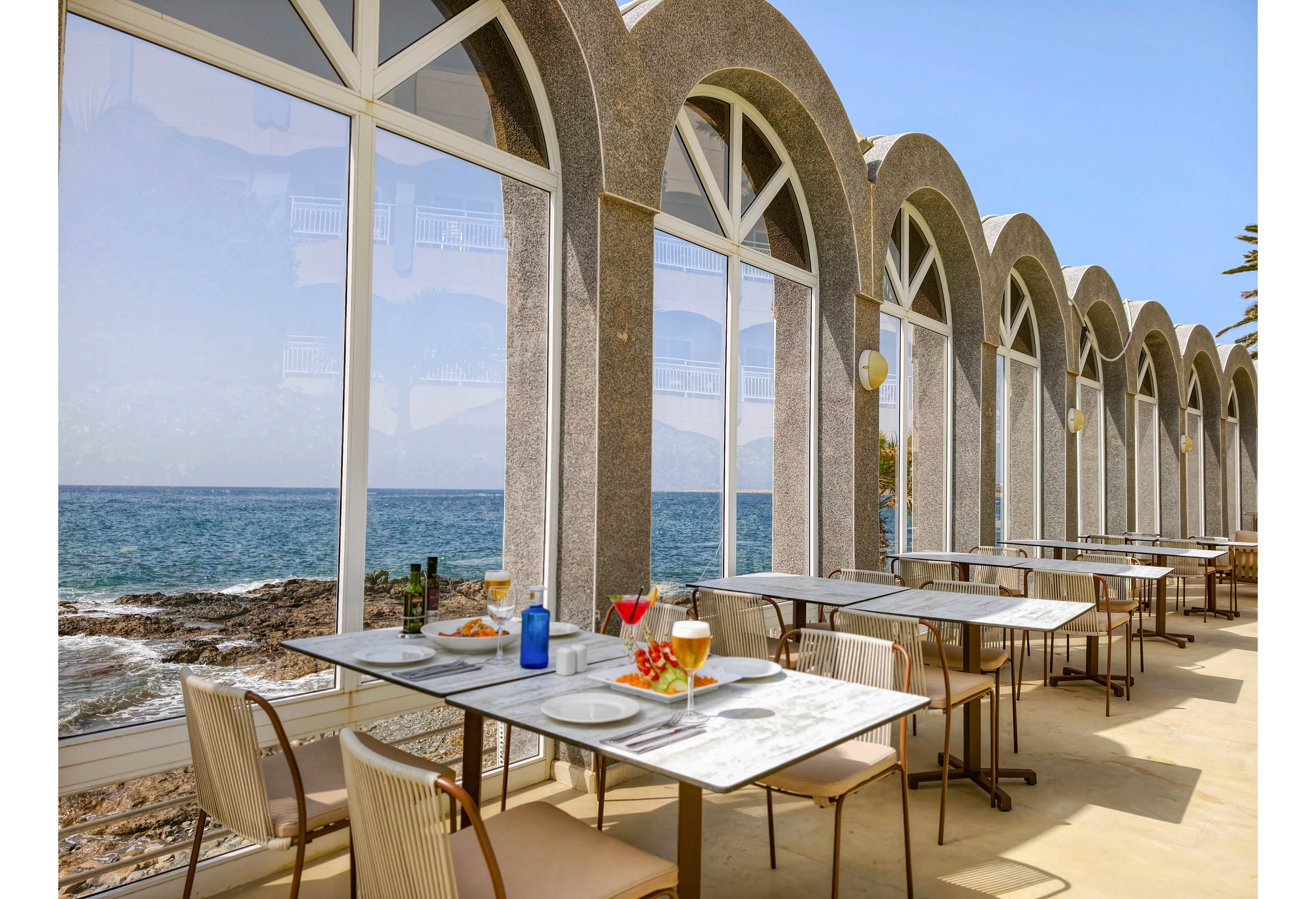 An elegant seaside restaurant terrace features marble-topped tables set for a meal beside large arched windows. The view overlooks a rocky shoreline and calm blue sea under a clear sky. Colorful drinks and plated dishes are arranged on the nearest table, suggesting a relaxed dining experience. Empty chairs and neatly aligned tables create an inviting yet tranquil atmosphere.