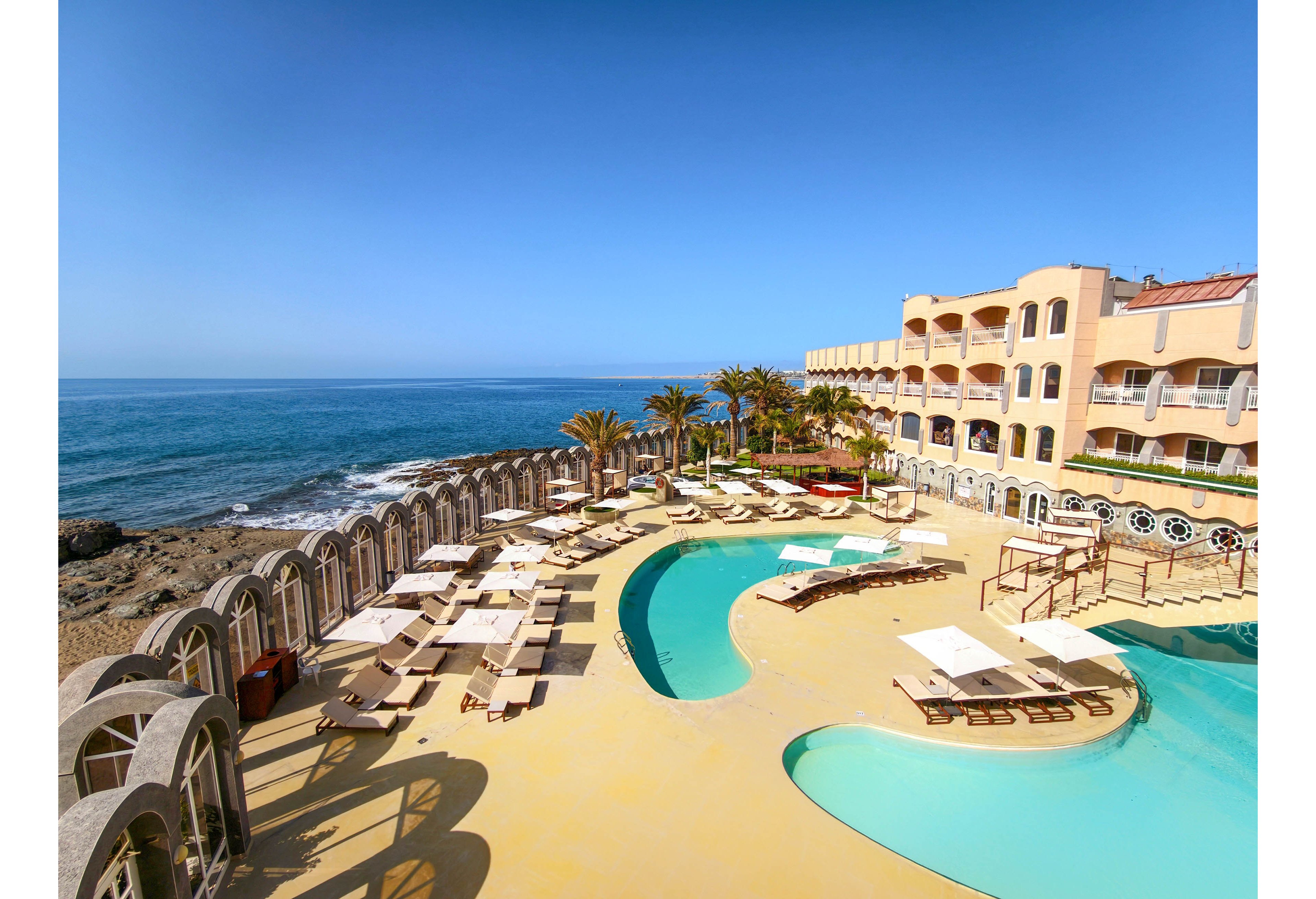 An oceanfront resort features a bright pool terrace overlooking a calm blue sea. Curved swimming pools sit at the center, surrounded by sun loungers, umbrellas, and arched stone railings. A multi-story hotel building with balconies faces the water, framed by palm trees and clear skies. The scene suggests a sunny, relaxing vacation destination on the coast.