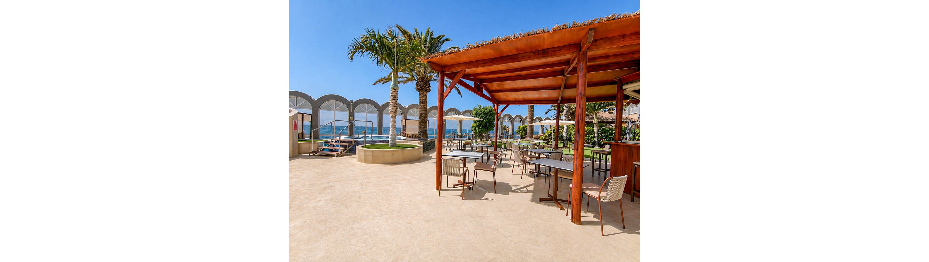 An open-air seaside terrace features a wooden pergola with tables and chairs arranged for outdoor dining. Palm trees and potted greenery add a tropical touch to the bright, sunlit setting. In the background, arched glass or acrylic panels frame views of the blue ocean and sky. The scene feels calm and inviting, ideal for relaxing, socializing, or enjoying a meal by the water.