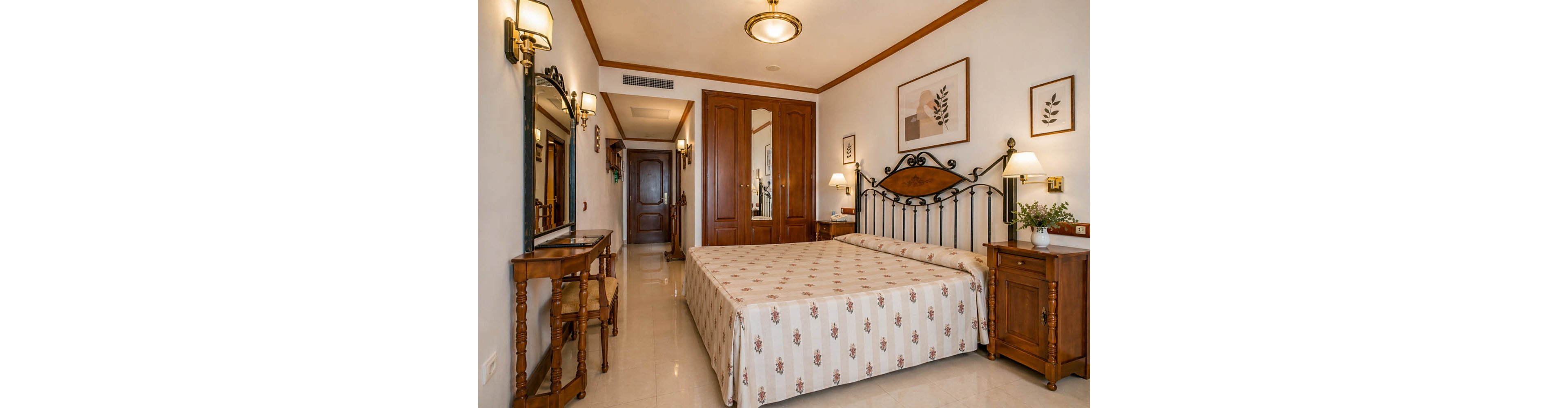 A warm, traditional hotel bedroom features a neatly made double bed with a patterned bedspread and an ornate metal headboard. Wooden furniture, including bedside tables, a writing desk, and a mirror, adds a classic touch to the room. Soft wall lighting and framed botanical artwork create a welcoming, homely atmosphere. The space appears clean, organized, and inviting for guests.