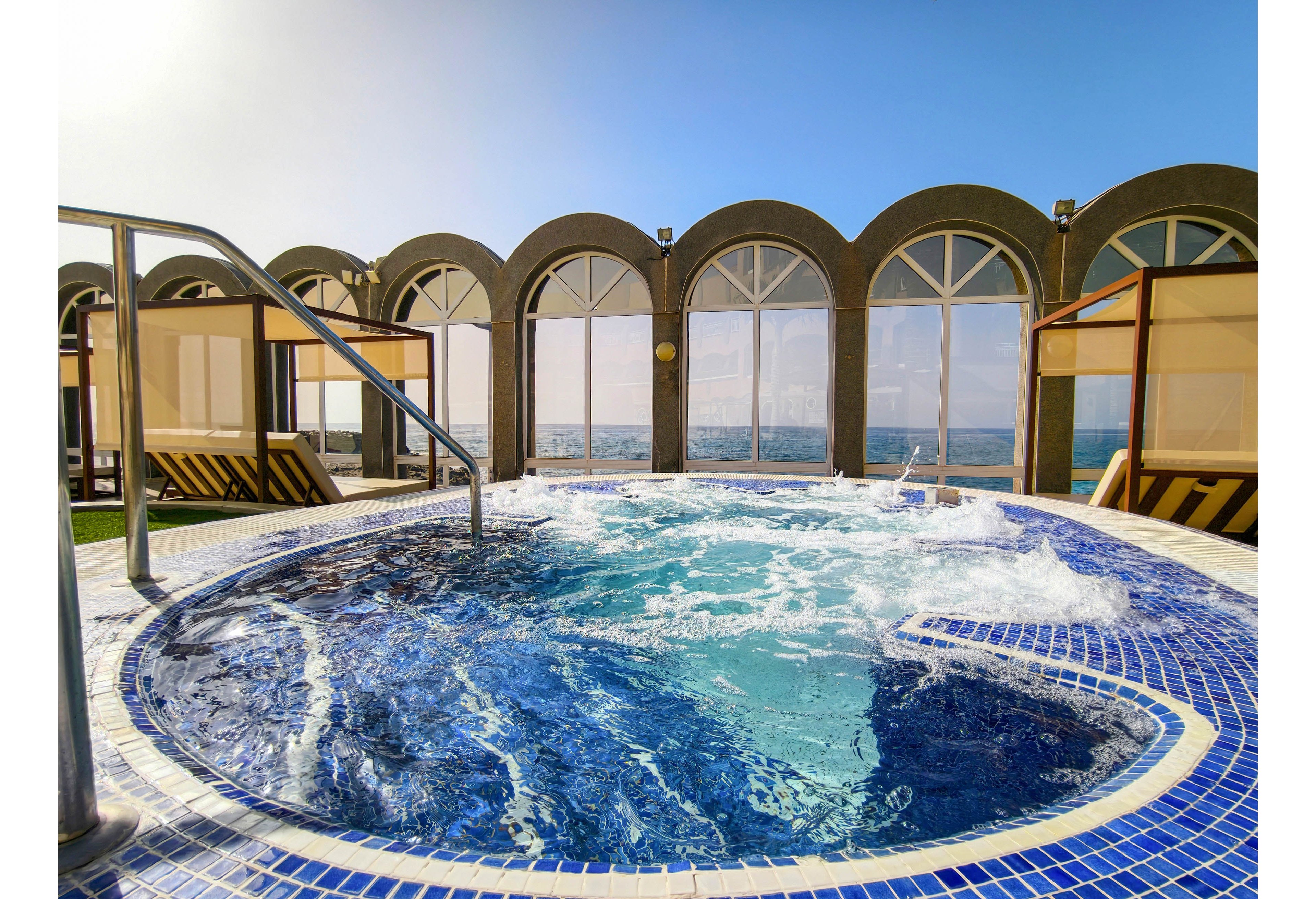 A circular jacuzzi with bubbling water sits on an outdoor rooftop deck. The pool is lined with blue and white tiles and features a metal handrail for easy access. Large arched windows and partitions frame expansive views of the ocean and clear sky. The bright daylight and calm seascape create a relaxing, luxurious spa atmosphere.