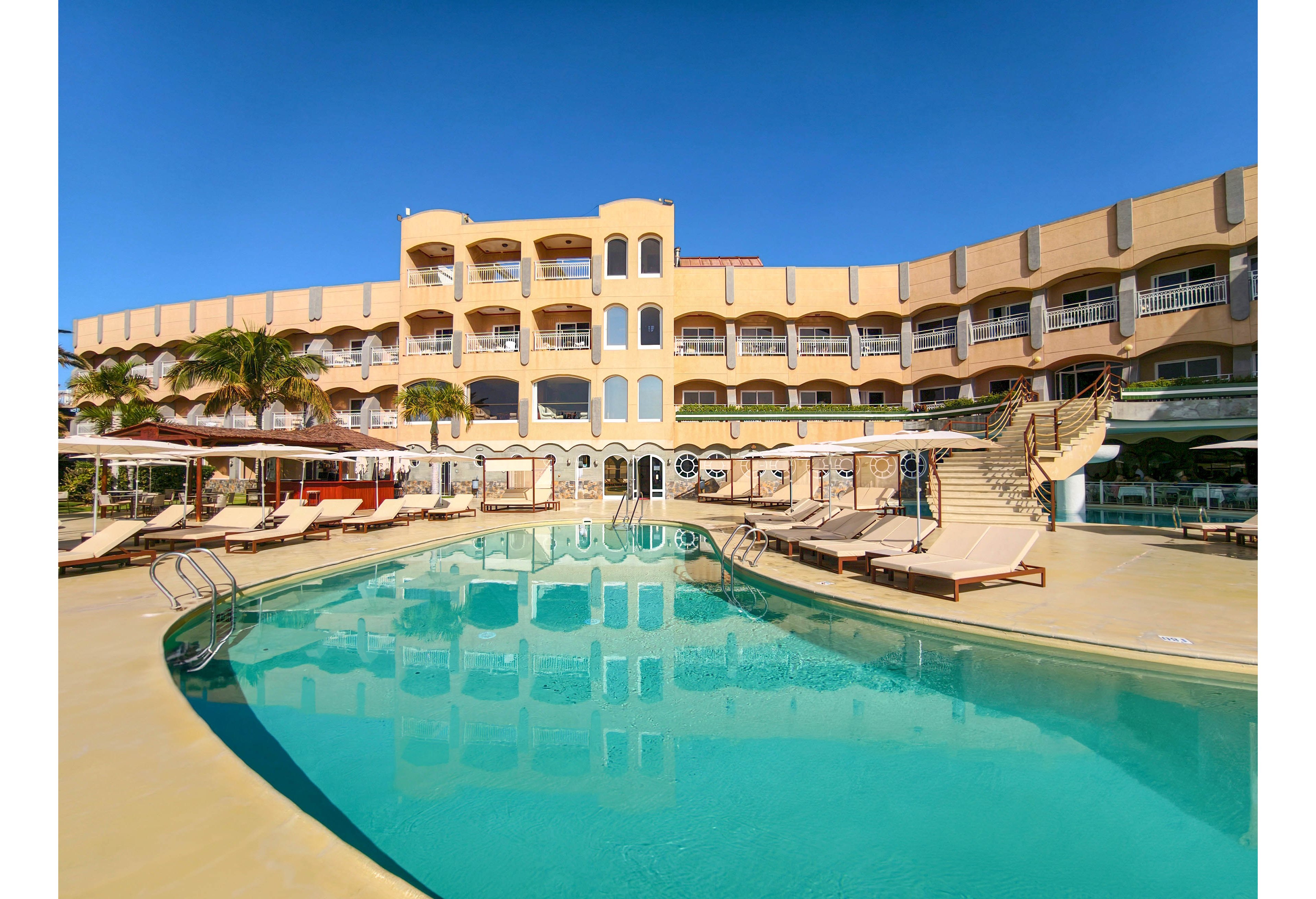 A large resort hotel features an expansive outdoor swimming pool in the foreground. Sun loungers and umbrellas line the poolside deck, suggesting a relaxing vacation setting. The multi-story building has arched windows and balconies overlooking the water. Clear blue sky and palm trees enhance the tropical, sunny atmosphere.