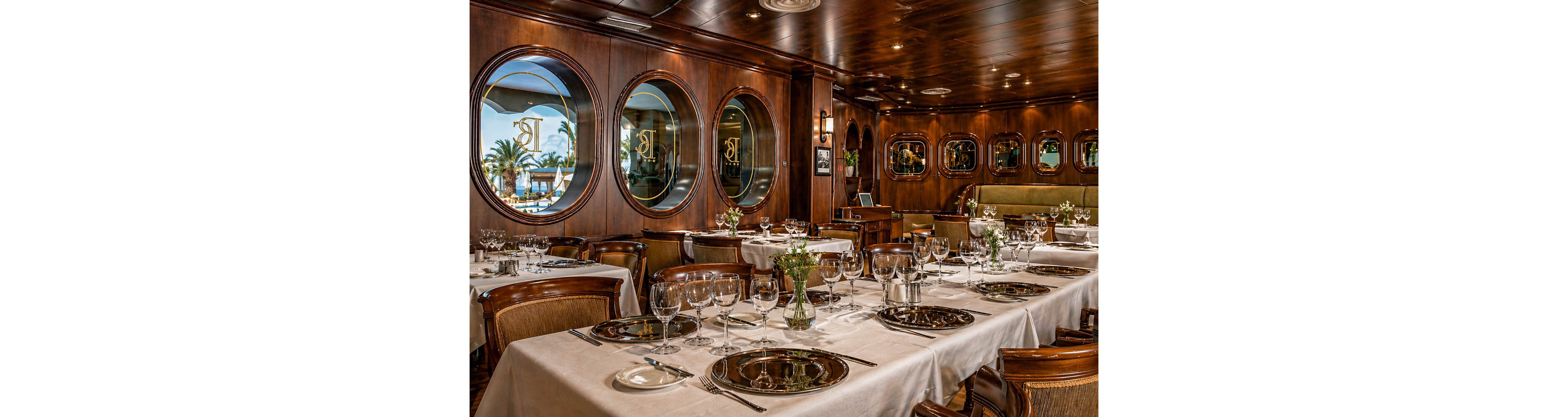 Upscale dining tables are set inside a wood-paneled restaurant that resembles a classic cruise ship interior. White tablecloths, polished glassware, and neatly arranged silverware create a formal atmosphere. Round porthole-style windows reveal palm trees and a bright waterfront outside. Soft lighting and small floral centerpieces add to the refined, intimate ambiance.
