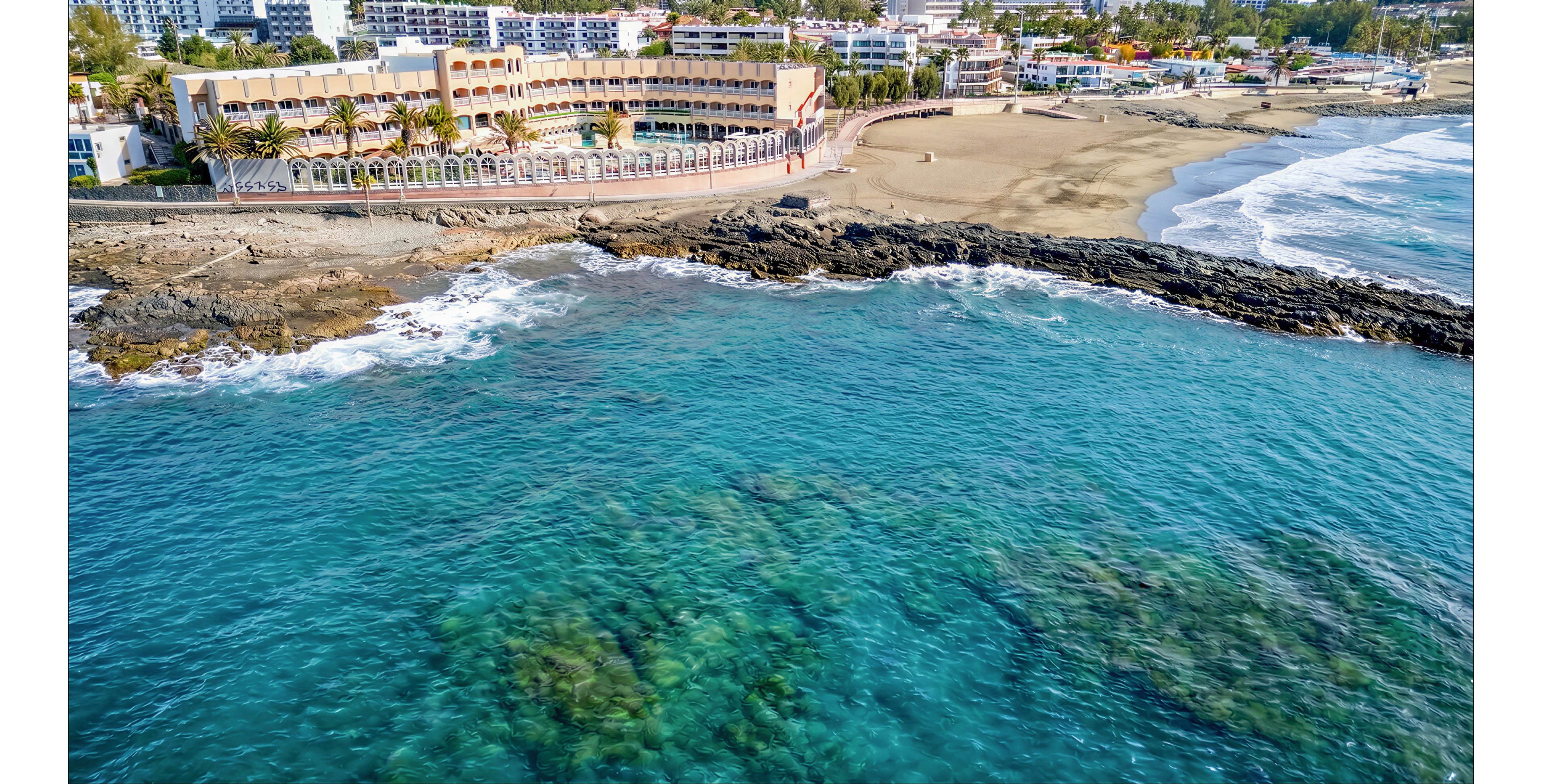 A seaside resort town stretches along a sandy beach beside clear turquoise water. The aerial perspective shows hotels and apartments lining the shore with palm trees and a promenade. Dark rocky outcrops separate the open ocean from the calmer bay area. Gentle waves roll toward the beach, and the water reveals submerged rocks and reefs near the coastline.