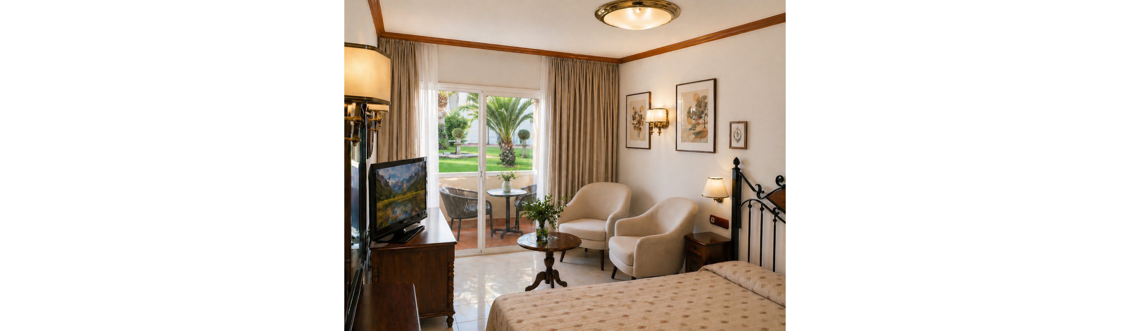 A warmly lit hotel room features a double bed, two upholstered armchairs, and a small round wooden table. A flat-screen television sits on a wooden cabinet opposite the bed, displaying a scenic landscape. Framed artwork and classic lamps add decorative touches to the neutral interior. Large glass doors open to a terrace with a table and chairs, overlooking a lush garden with palm trees.
