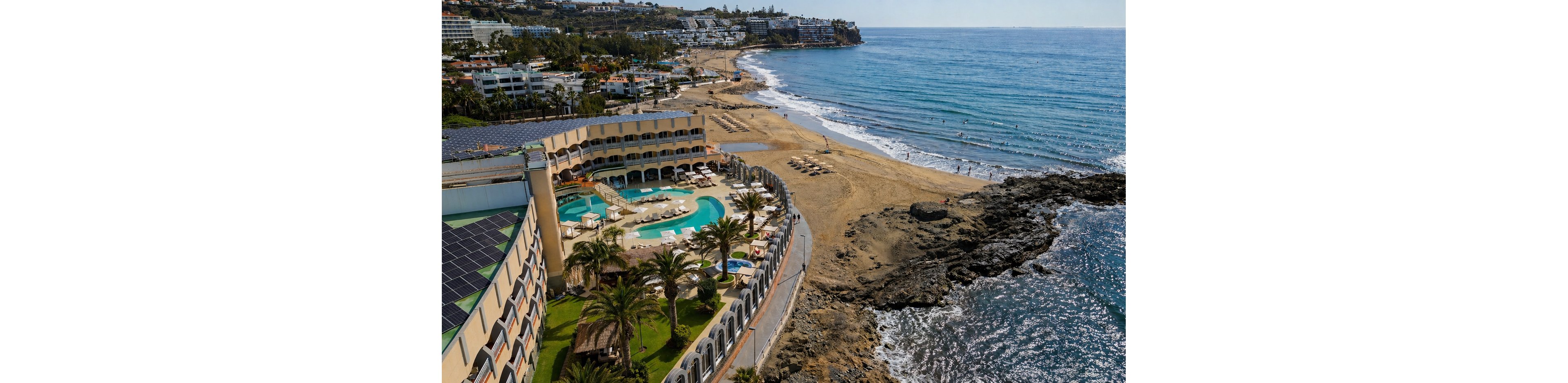 A large coastal resort curves along the shoreline beside a wide sandy beach. The hotel features an outdoor swimming pool, palm trees, and sun loungers arranged near the water. Rocky formations border part of the coast while gentle waves roll in from the ocean. In the distance, additional buildings and greenery line the waterfront under a clear sky.