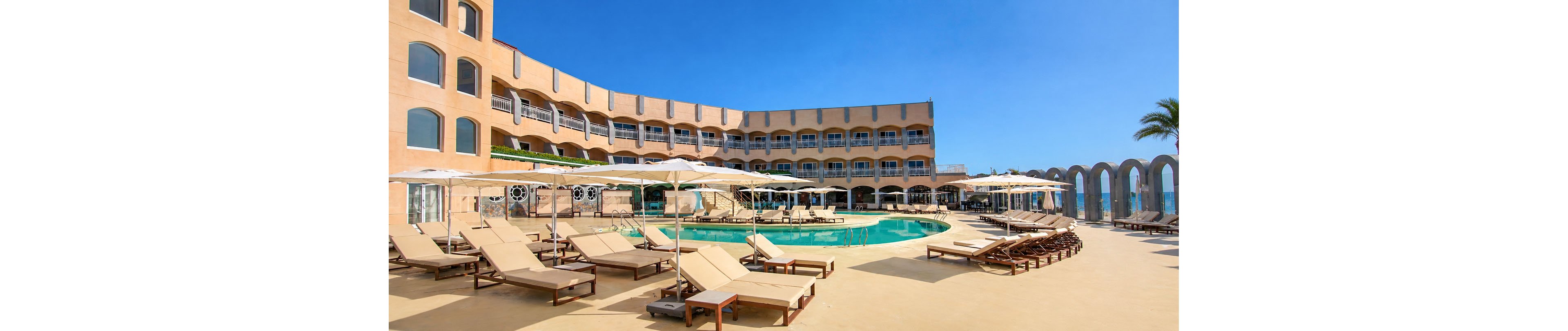A curved resort building surrounds an outdoor swimming pool under a clear blue sky. Numerous beige lounge chairs and white sun umbrellas line the poolside deck, suggesting a relaxing vacation setting. The architecture features arched windows and decorative arches, with a palm tree visible on the right side. Bright sunlight and warm tones create a tranquil, inviting atmosphere.