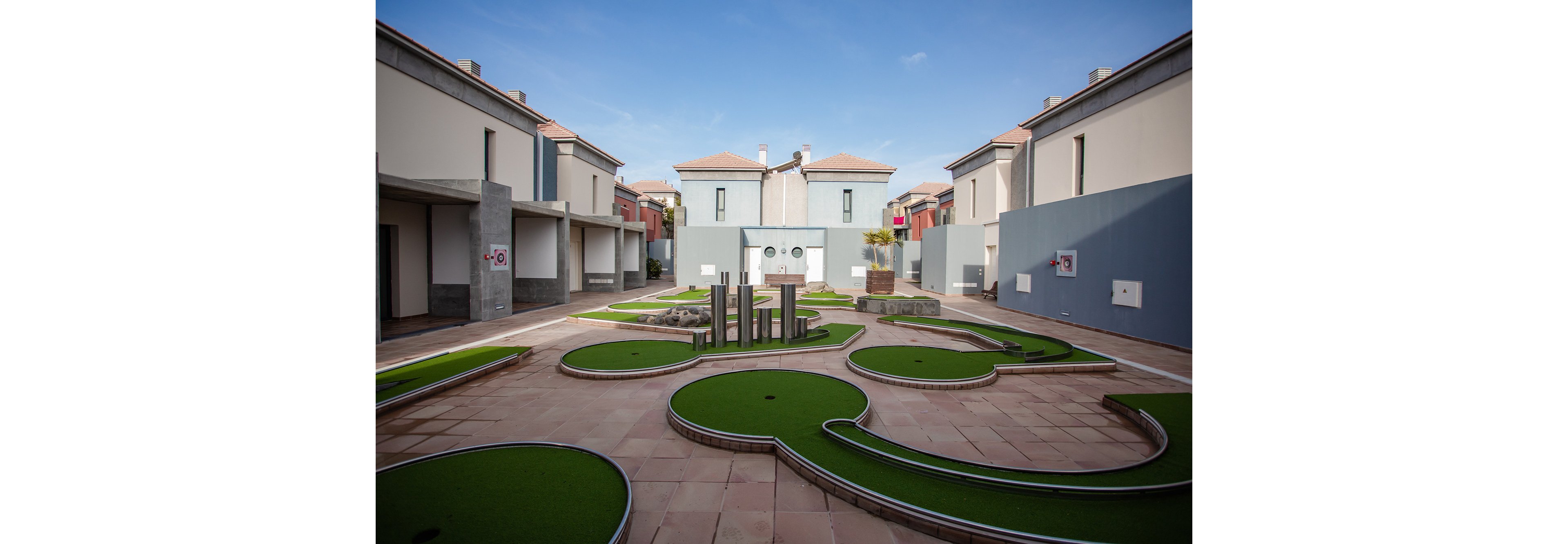 A contemporary mini golf course is set in the courtyard between rows of modern townhouses. Curved putting greens with artificial turf are surrounded by smooth stone or tile walkways. Simple geometric obstacles and low metal poles add visual interest to the course. The sky is clear and blue, giving the scene a bright, relaxed atmosphere.