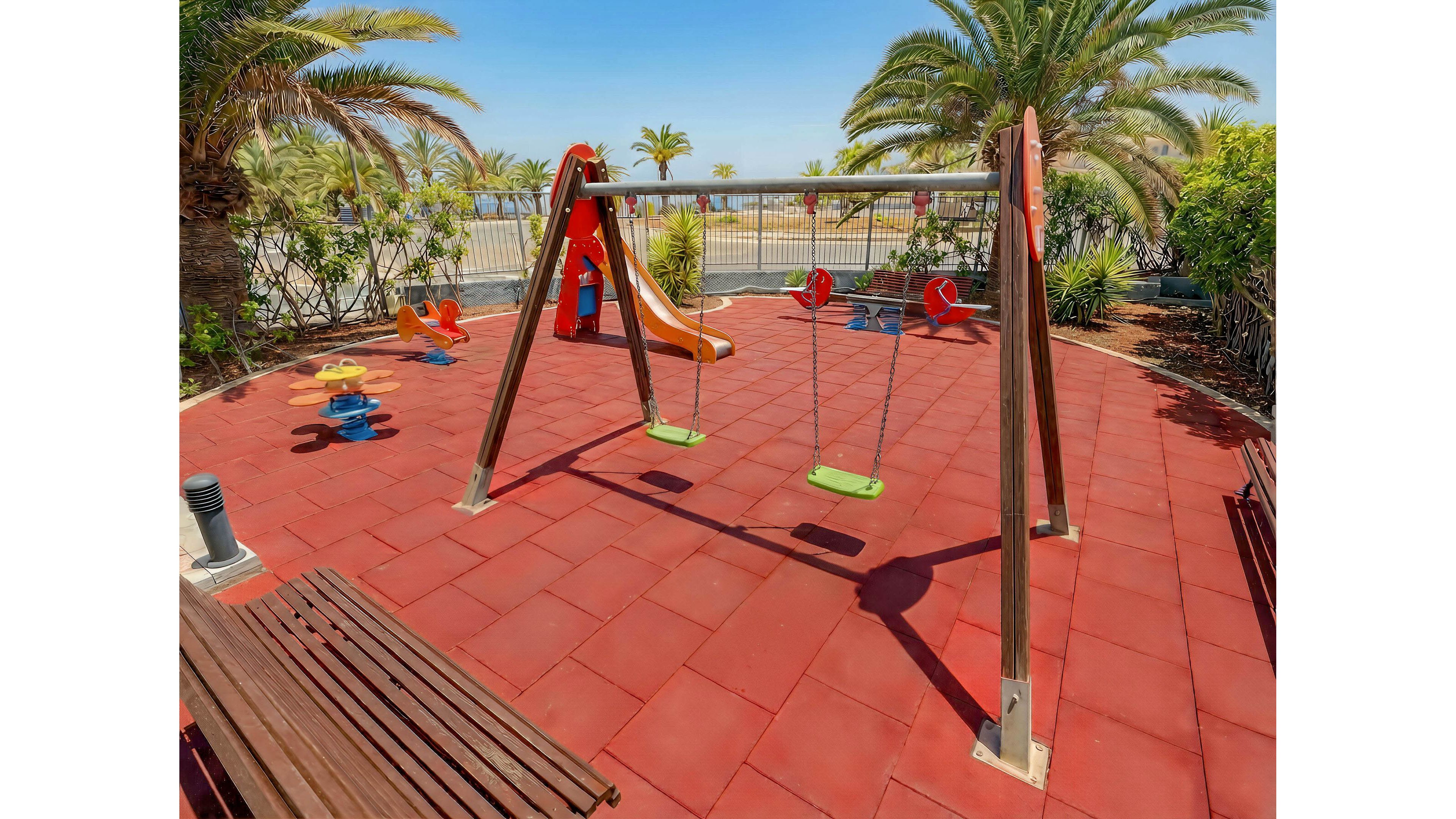 An empty children's playground features a swing set and a small slide on a red rubber safety surface. Colorful spring riders are scattered around the play area, which is bordered by lush green bushes and palm trees. In the background, a metal fence separates the playground from a seaside promenade with more palm trees and a clear blue sky. The bright daylight and vivid colors create a cheerful, inviting atmosphere.