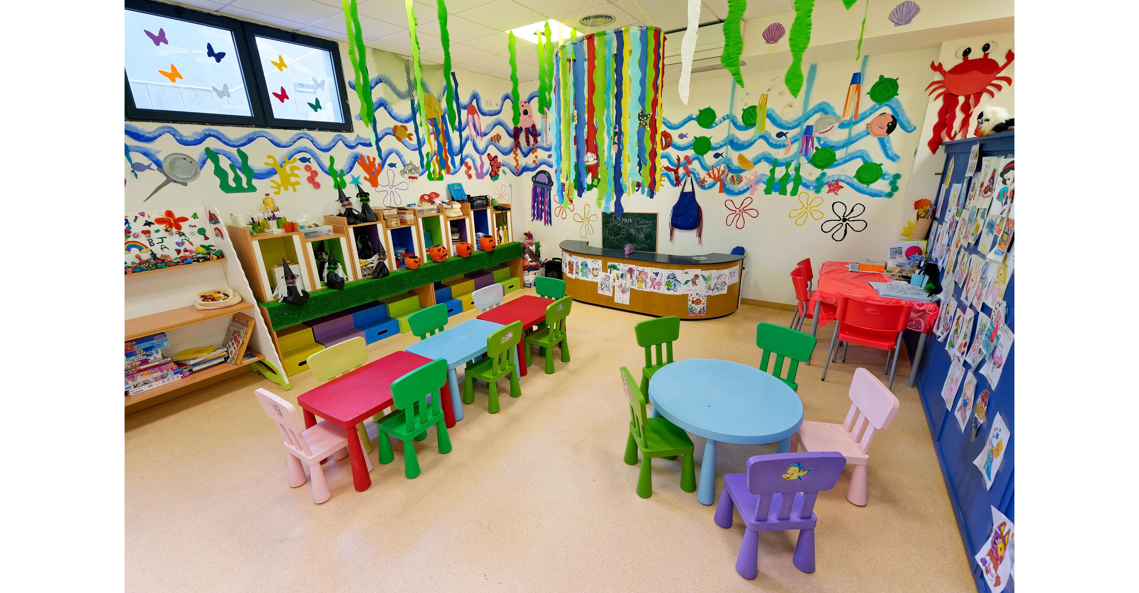 A bright preschool classroom is filled with small colorful tables and chairs arranged for young children. The walls are decorated with playful murals of waves, sea creatures, butterflies, and flowers. Hanging streamers and paper decorations add to the festive, creative atmosphere. Shelves hold books, toys, and art supplies, suggesting a space designed for learning and imaginative play.