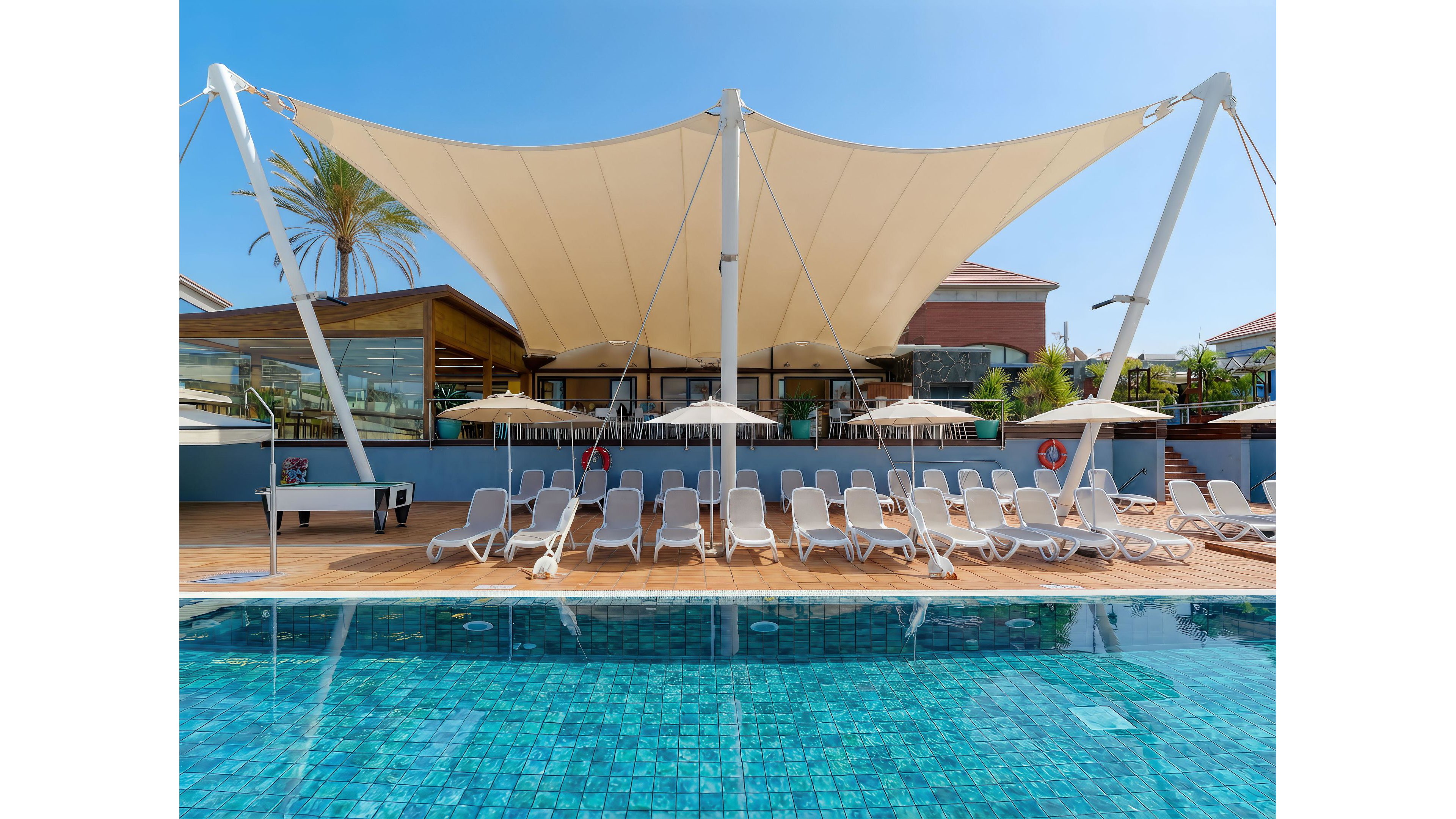 A row of empty white sun loungers lines a wooden deck beside a clear blue swimming pool. A large tensile shade canopy stretches overhead, providing shelter from the bright sun. Beige parasols and a modern resort building with glass walls appear in the background. The scene suggests a calm, sunny vacation atmosphere at a contemporary pool area.