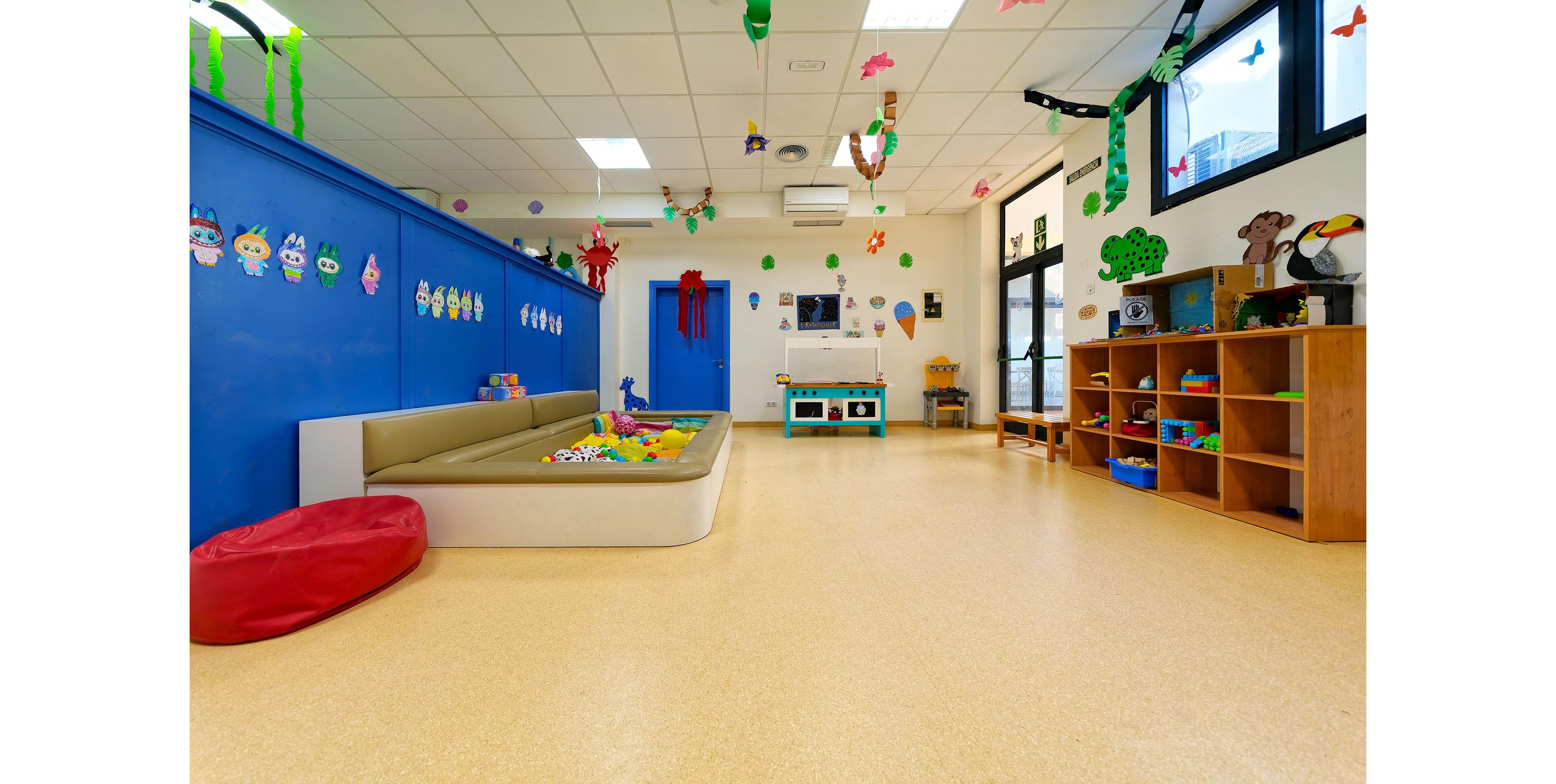A bright kindergarten classroom features a large open play area with a cushioned pit filled with toys. Blue walls are decorated with cheerful cartoon characters and hanging crafts. Wooden shelves on the right hold books, games, and educational materials. Large windows let in natural light, creating a welcoming and safe environment for young children.
