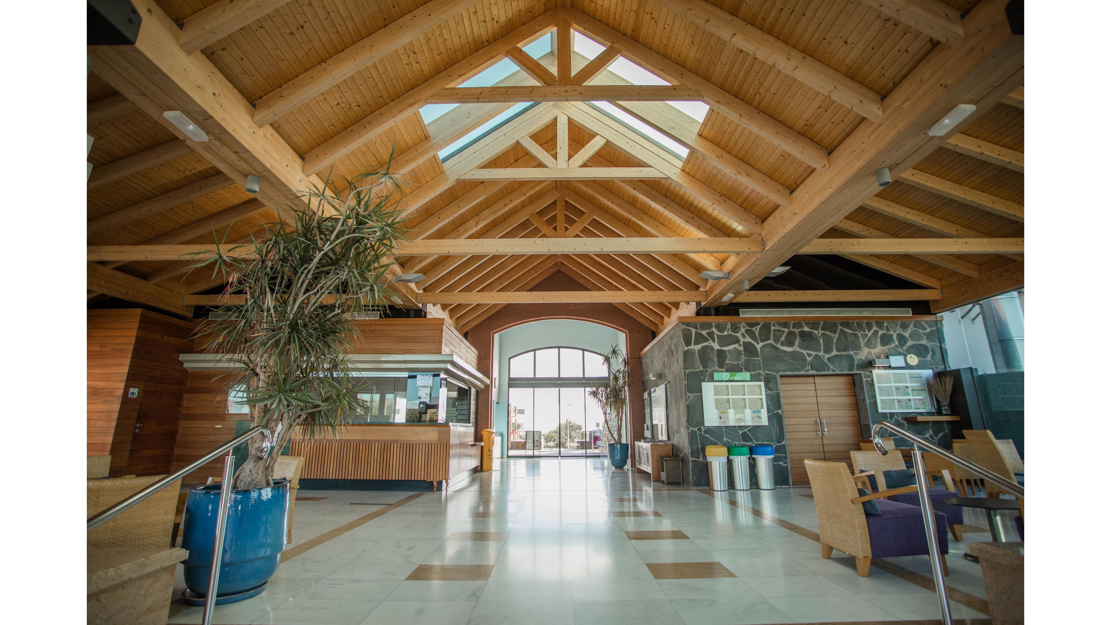 A bright, spacious lobby features a dramatic exposed wooden beam ceiling and large skylight. The interior includes a reception or bar counter on the left and stone accent walls on the right. Natural light floods in through a large glass doorway at the far end, highlighting polished floors and scattered seating. Potted plants and colorful bins add visual interest and a welcoming atmosphere.