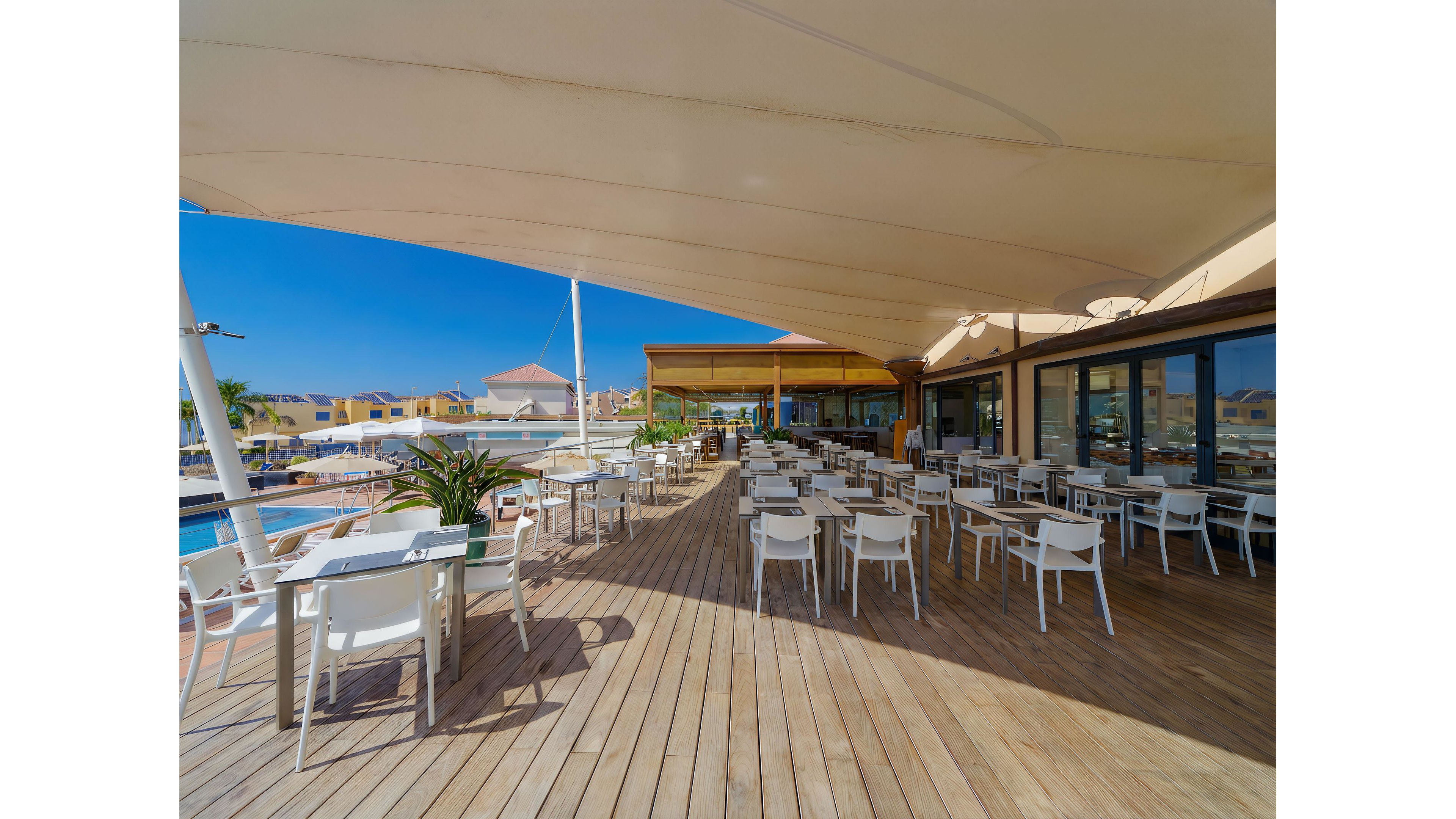 An open-air restaurant terrace features rows of white tables and chairs on a wooden deck. A large fabric canopy provides shade over the seating area, while glass doors lead into the interior space. In the background, a swimming pool and resort buildings are visible under a clear blue sky. The scene suggests a relaxed, sunny vacation atmosphere at a modern resort.