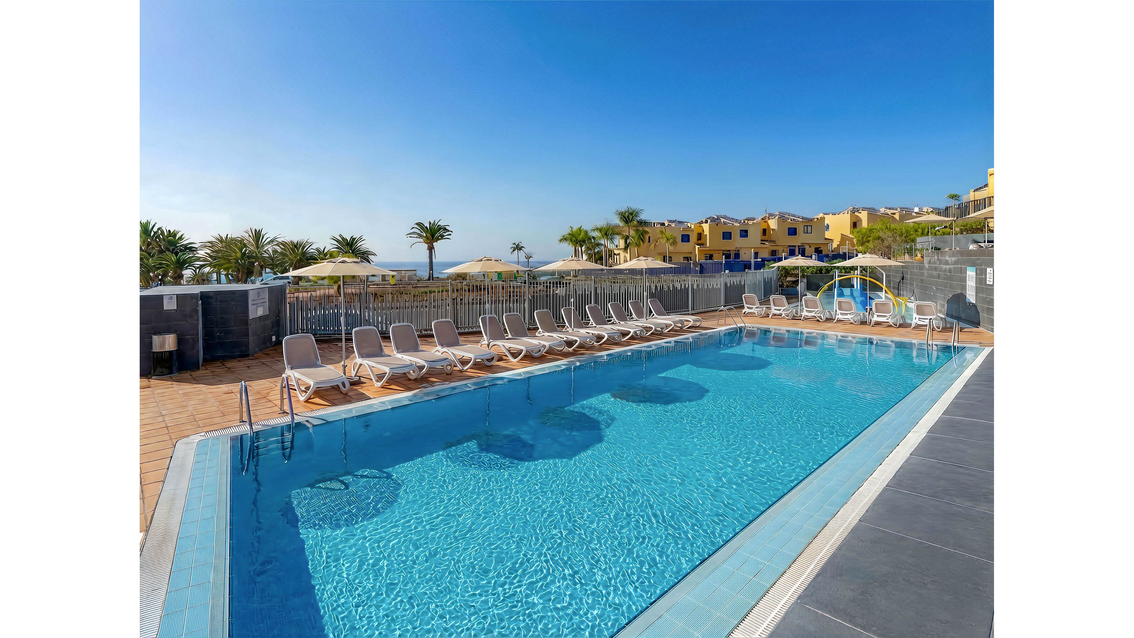 An outdoor swimming pool glistens under a clear blue sky at a resort. Numerous white sun loungers line the poolside, all facing the water and neatly arranged in rows. Yellow buildings and palm trees are visible in the background, suggesting a warm coastal or vacation destination. The scene feels calm and inviting, ideal for relaxation and leisure by the pool.
