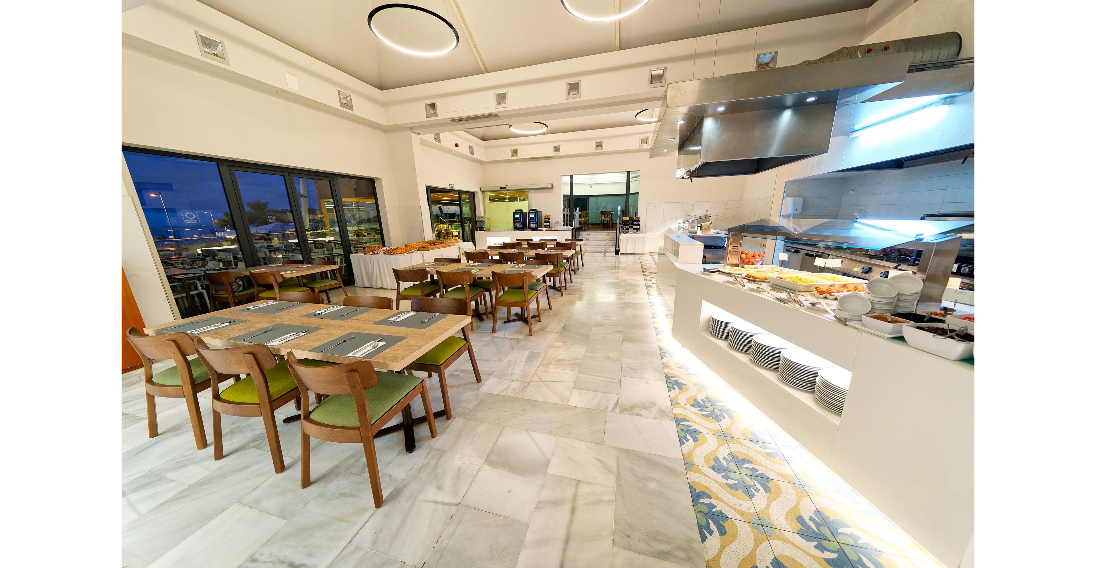 A bright, modern restaurant dining area features neatly arranged wooden tables and chairs with green cushions. An open buffet counter on the right displays various dishes and plates under stainless steel hoods. Large windows and glass doors reveal an evening view outside, while ceiling lights provide warm illumination. The clean marble floor and patterned tile strip add a contemporary, inviting atmosphere.