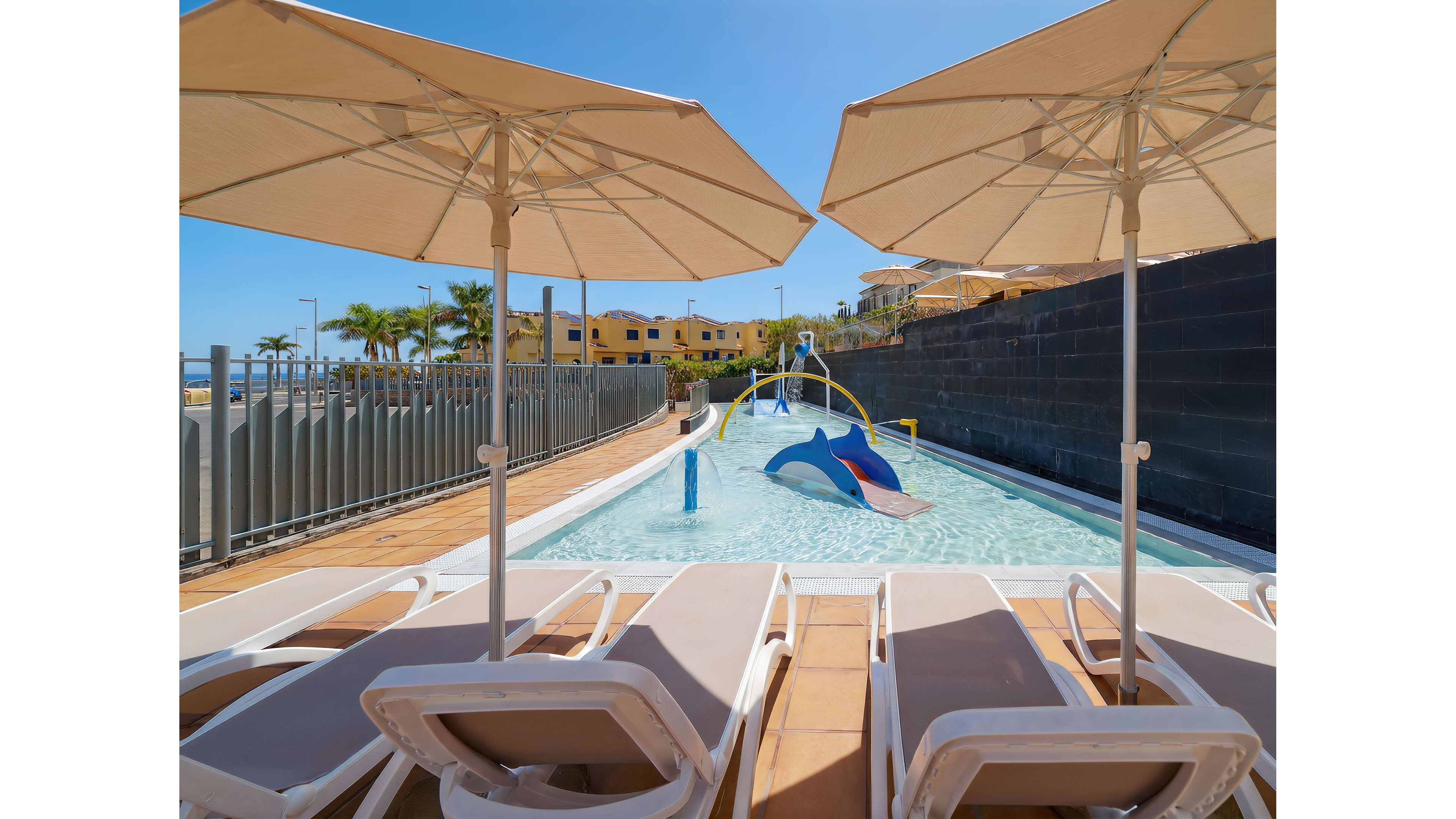 A small shallow pool with playful dolphin and water-spray structures sits in the center of a sunny resort area. Several lounge chairs with cushions are arranged under large white umbrellas along the poolside. In the background, palm trees and low yellow buildings suggest a warm vacation destination. The clear blue sky and bright sunlight create a relaxed, family-friendly atmosphere.