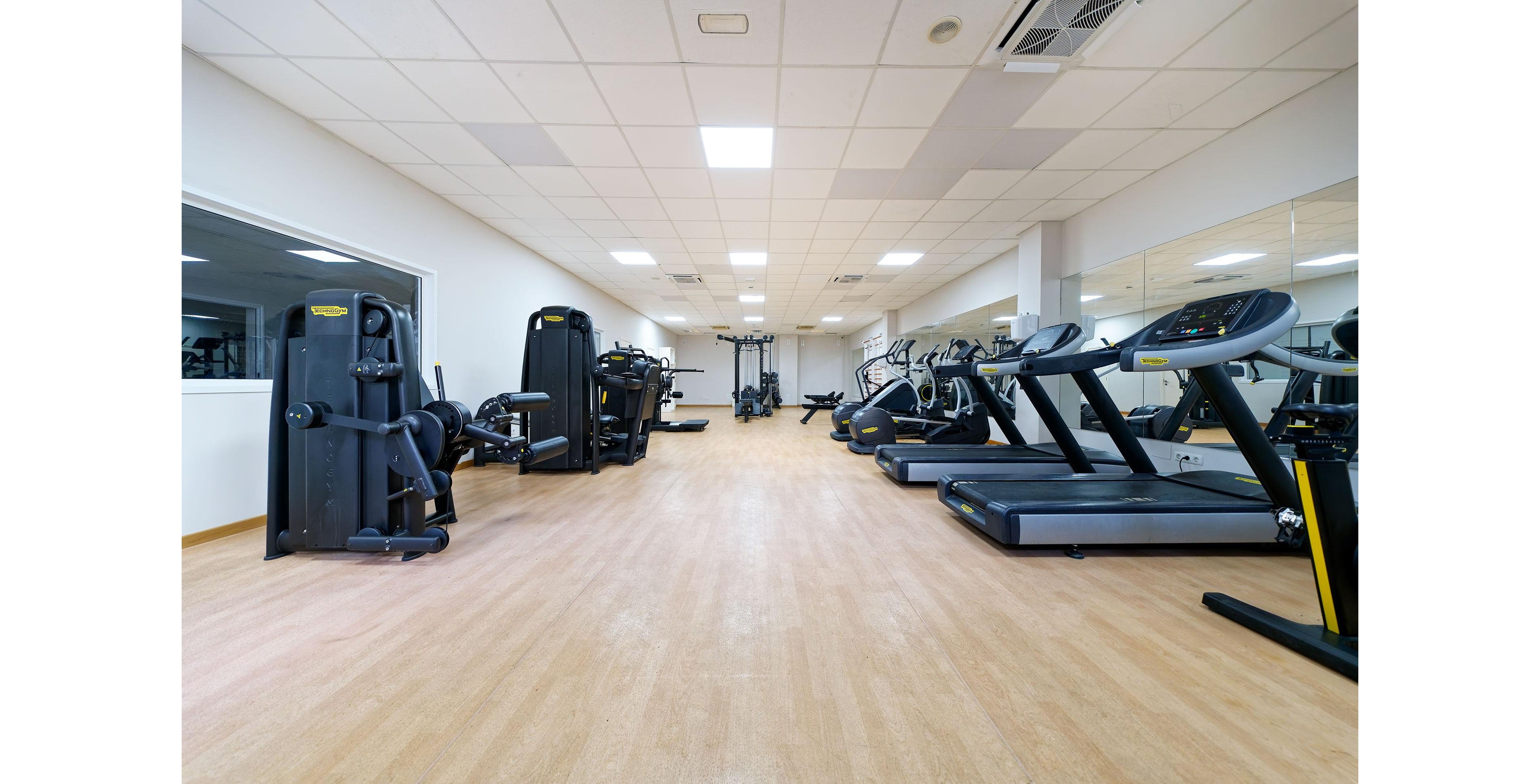 A bright, modern gym interior features rows of treadmills and strength machines neatly arranged along the walls. The room has a light wood-style floor, white walls, and a suspended ceiling with recessed lighting. Large mirrors line one side of the space, visually expanding the room and reflecting the equipment. The gym is empty, creating a clean, organized, and spacious atmosphere suitable for fitness and training.
