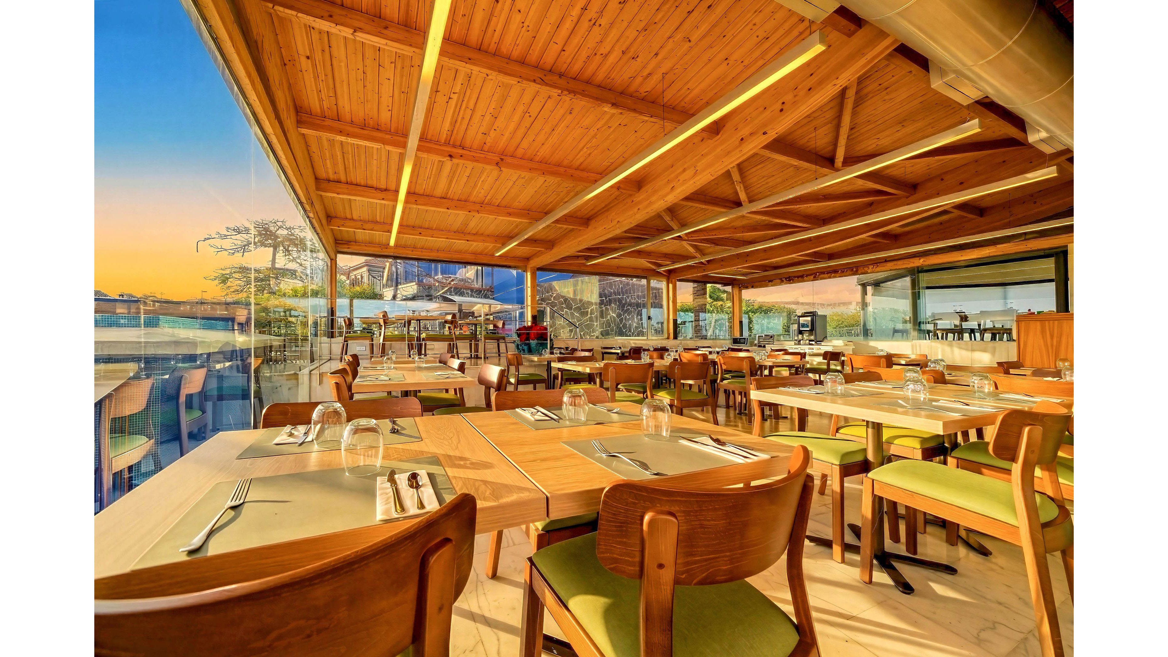 A modern restaurant dining area features neatly arranged wooden tables and chairs under a warm wooden ceiling. Large glass walls open to a scenic outdoor view with a colorful sunset sky. Place settings with plates, glasses, and cutlery are prepared for guests, but the space is currently empty. The overall atmosphere feels inviting, bright, and relaxed, ideal for evening dining with a view.