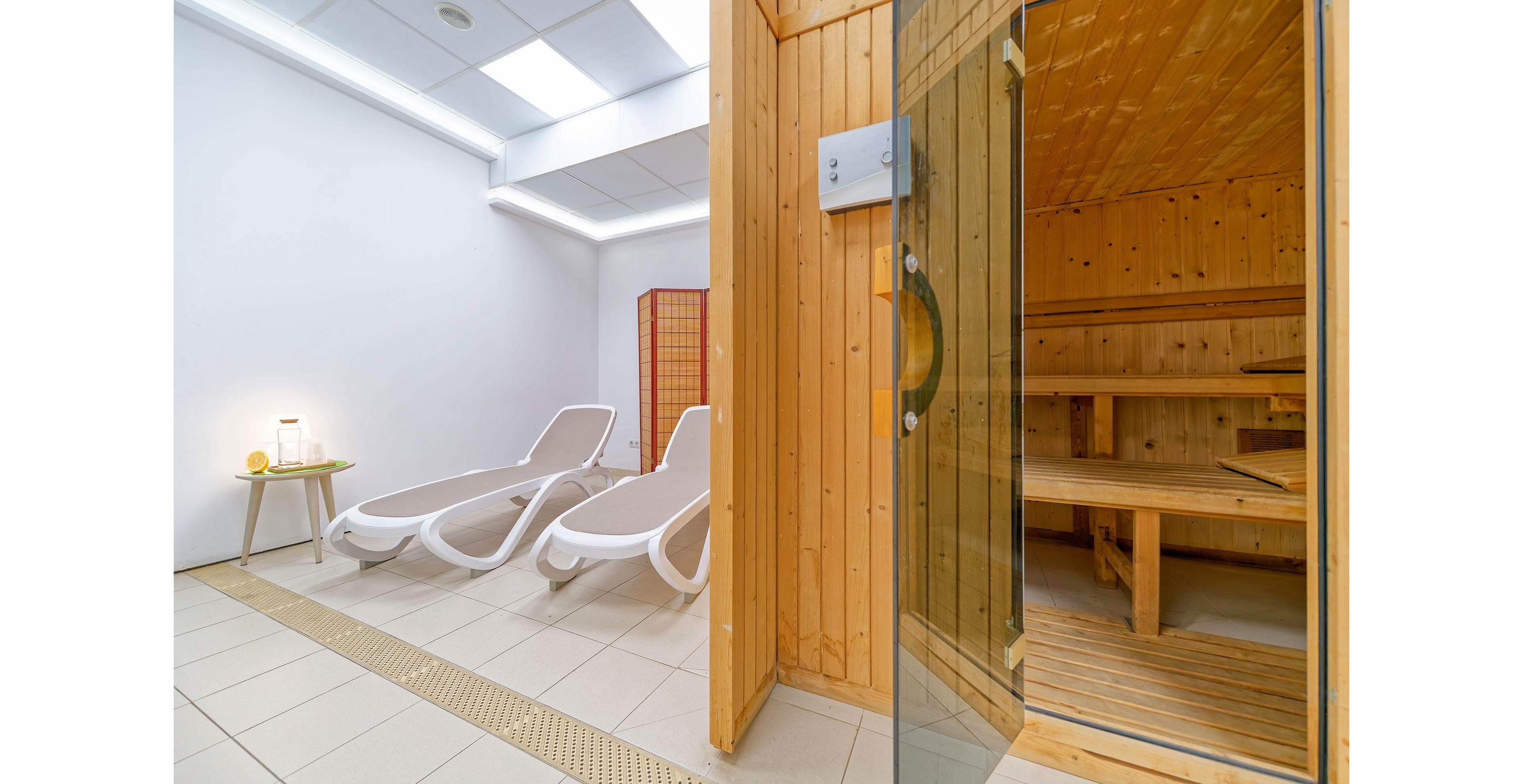 A contemporary spa area features a wooden sauna with a glass door on the right side. Next to the sauna, two white reclining lounge chairs are arranged in a bright, tiled relaxation room. The space is illuminated by ceiling lights and includes a small side table with a lamp and accessories. Clean lines, light wood, and neutral colors create a calm, minimalist atmosphere.