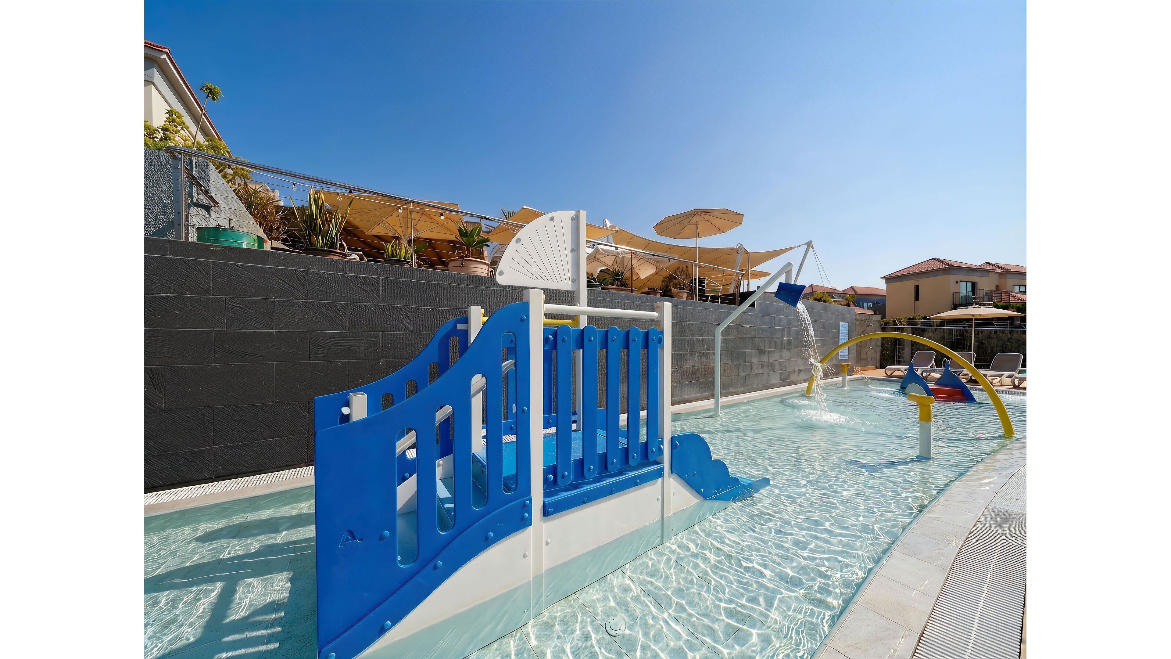 A shallow outdoor pool features a colorful children’s play structure with blue and white slides and railings. Water sprayers and curved pipes create playful fountains along the pool. Sun umbrellas and potted plants line the elevated deck above the pool. Residential-style buildings and a clear blue sky complete the bright, family-friendly setting.