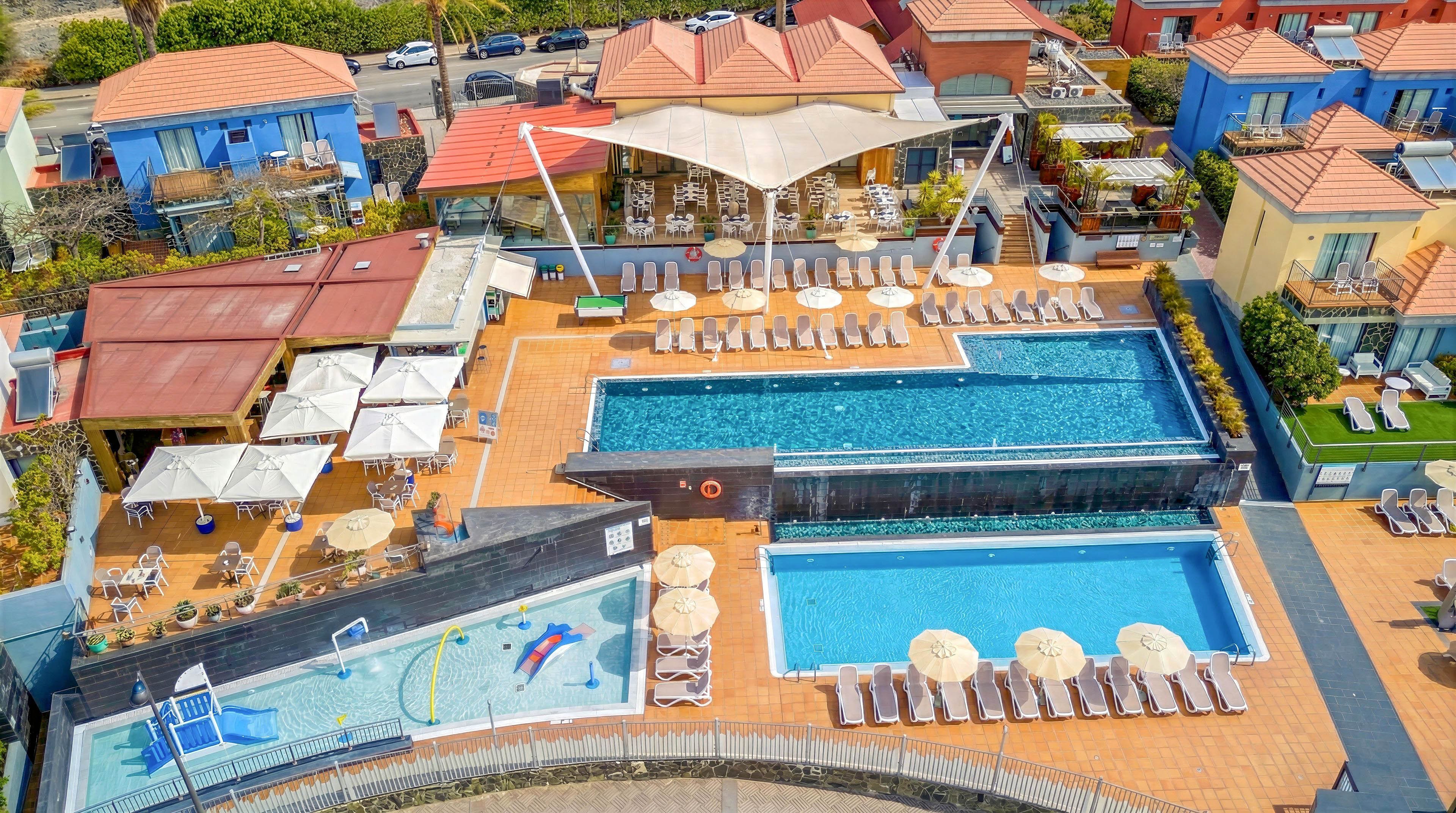 An aerial view shows a resort-style swimming pool complex with multiple pools and sun loungers. The main rectangular pool is surrounded by neatly arranged deck chairs and umbrellas on a terracotta-tiled terrace. A smaller pool and a shallow children's pool with a dolphin graphic are visible in the foreground. Colorful low-rise buildings with red roofs and landscaped paths frame the leisure area, creating a vibrant holiday atmosphere.