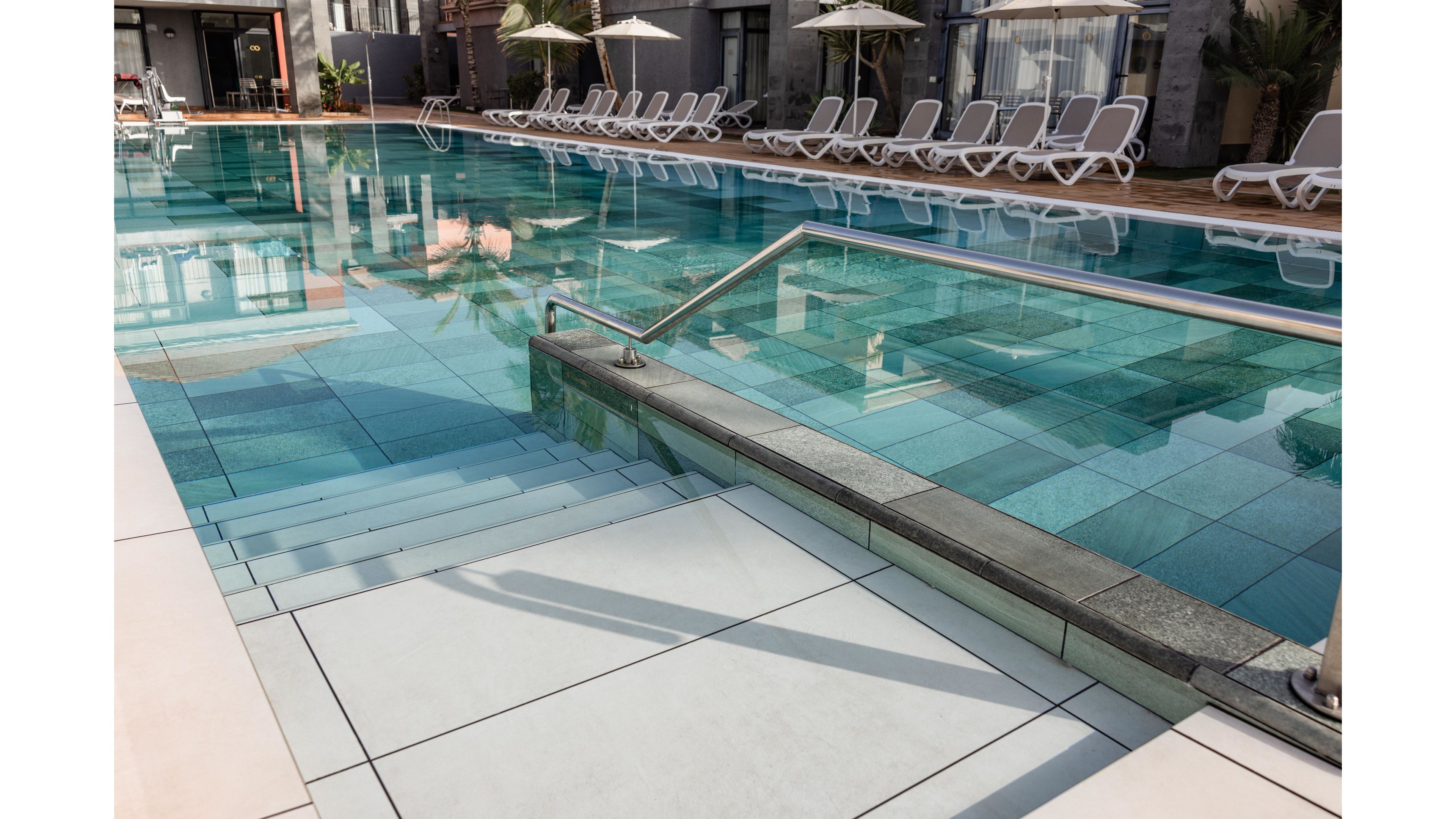A clear blue swimming pool sits in the courtyard of a modern resort or hotel. White lounge chairs and umbrellas line the far side of the pool, all currently unoccupied. Wide tiled steps lead into the shallow end, with a metal handrail providing safe access. The calm water and clean geometric tiles create a serene, inviting atmosphere for relaxation.