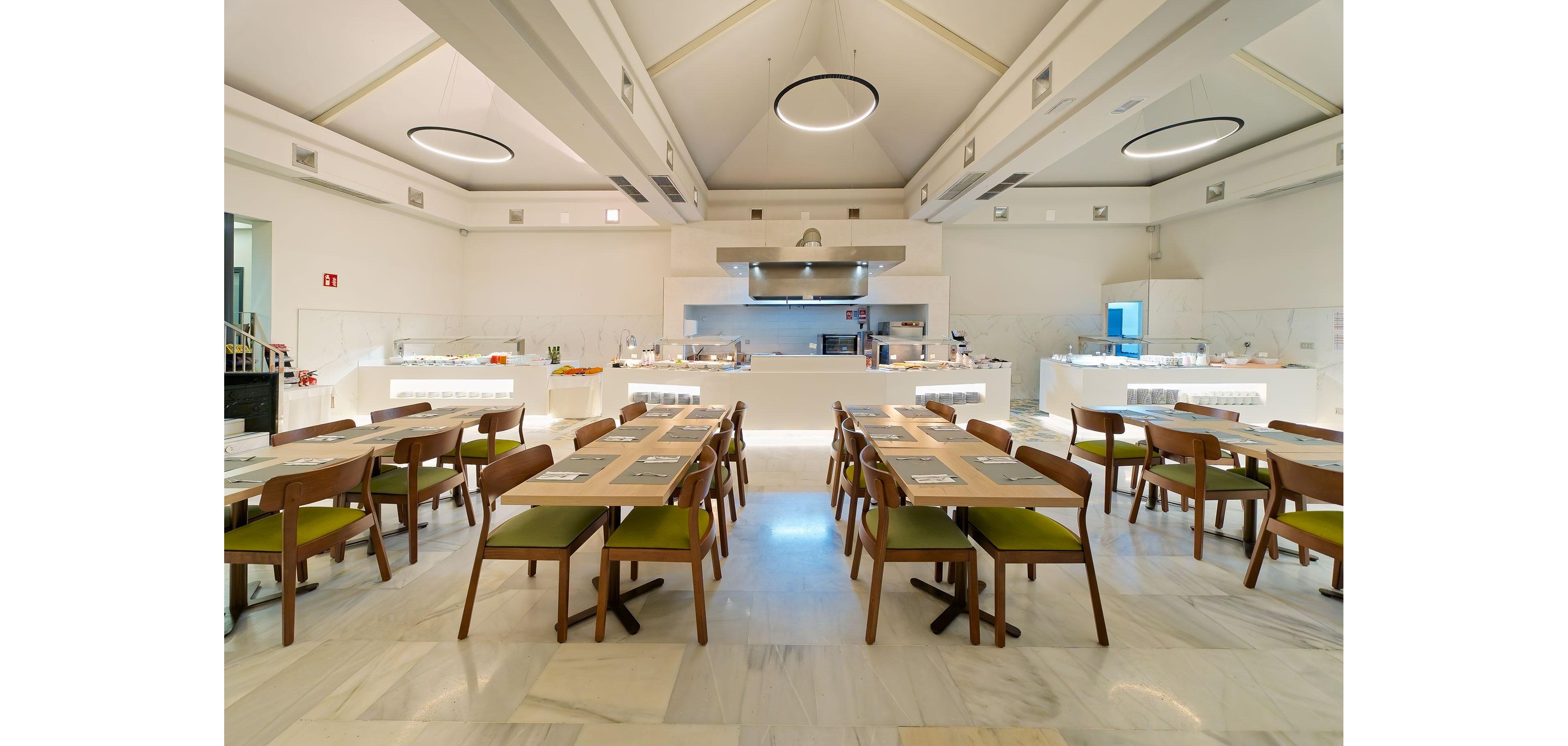 A bright, modern restaurant dining room features rows of wooden tables and chairs neatly arranged. The space is set up in a self-service style with buffet counters and a visible open kitchen at the back. Large circular ceiling lights and high vaulted ceilings create an airy, contemporary atmosphere. The room is empty of people, emphasizing the clean, organized layout and minimalist decor.