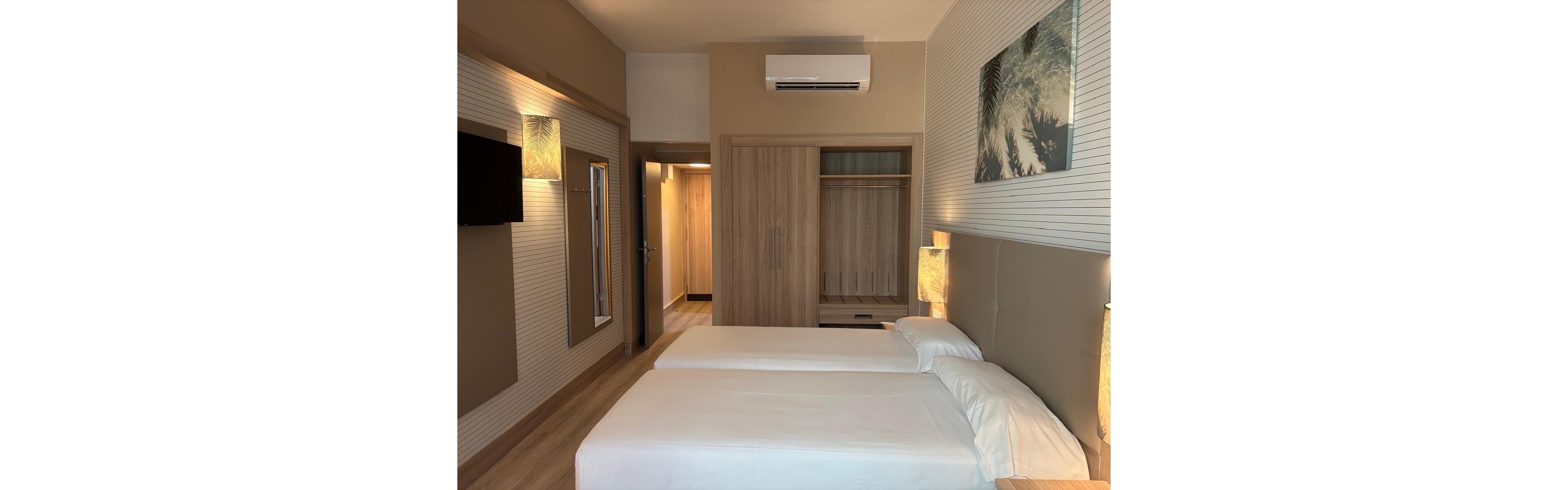 Contemporary hotel bedroom featuring two neatly made twin beds with white linens. The room includes a wooden wardrobe with open shelving and a doorway leading to a lit hallway. Warm bedside lamps, wall-mounted television, and decorative artwork create a cozy atmosphere. Neutral beige and wood tones dominate the interior, complemented by soft ambient lighting.