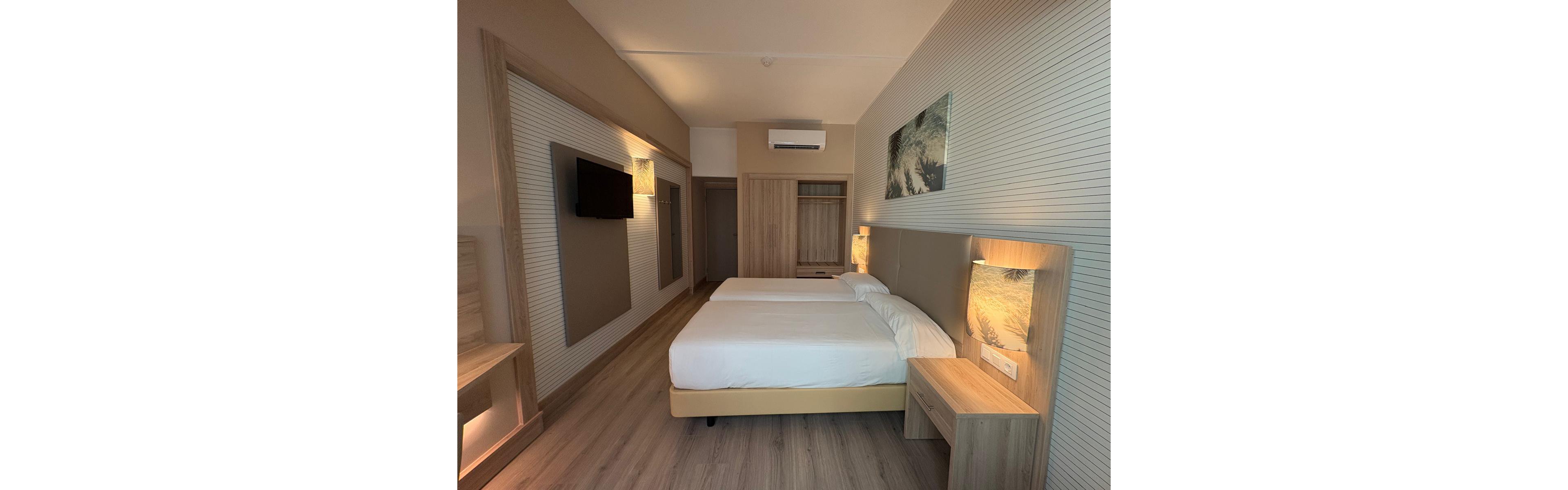Contemporary hotel room interior featuring two neatly made twin beds with upholstered headboards. The space includes light wood furniture, a wall-mounted flat-screen TV, and built-in wardrobe. Soft, warm lighting and neutral beige tones create a calm, minimalist atmosphere. A framed wall art piece above the bed adds a subtle decorative touch to the clean design.