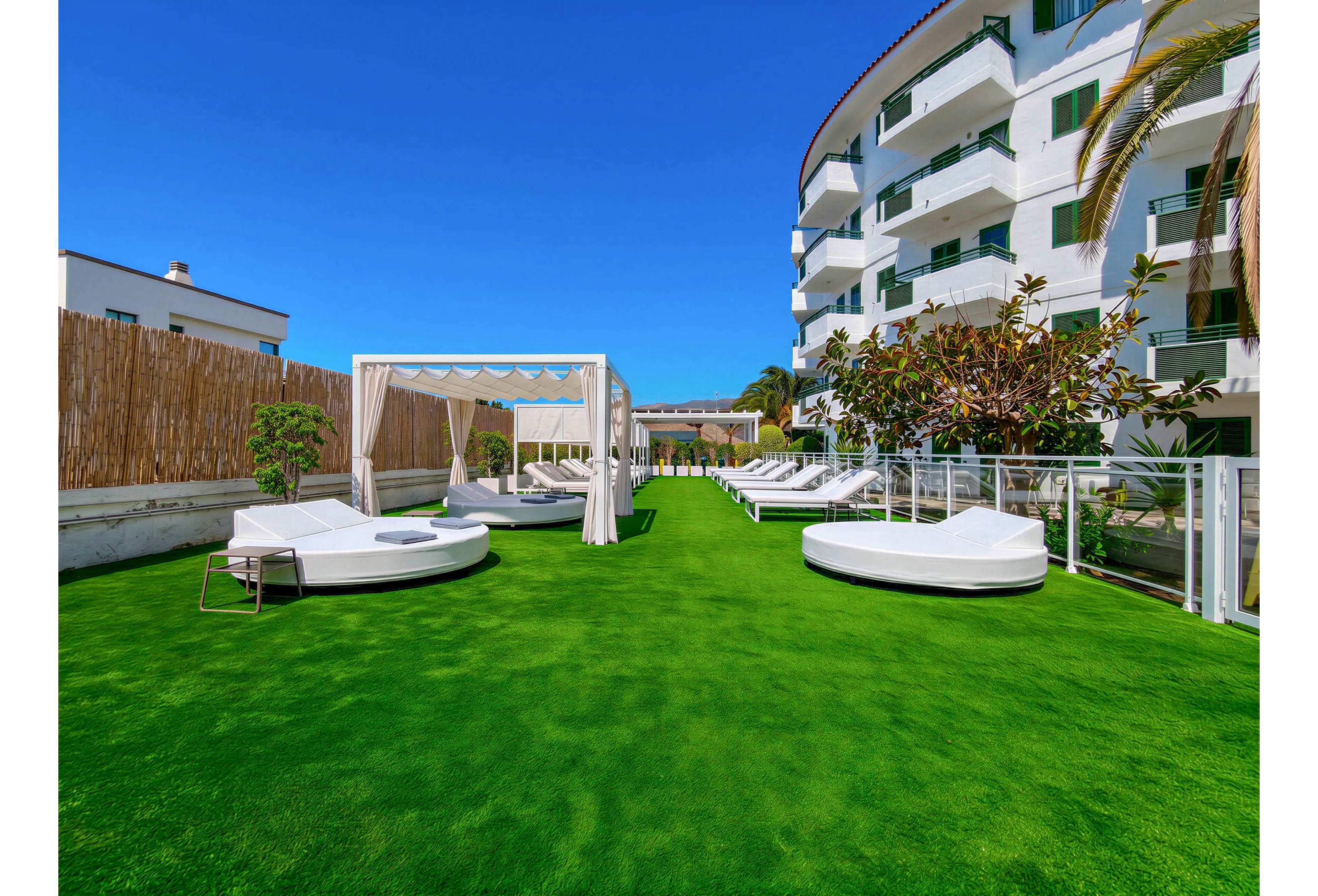 An outdoor hotel terrace features bright green artificial grass and modern white sun loungers. A canopy daybed sits to the left, with additional loungers arranged along glass railings and fencing. A multi-story white building with balconies and green shutters borders the space, under a clear blue sky. Palm trees and potted plants add a touch of greenery to the contemporary leisure setting.