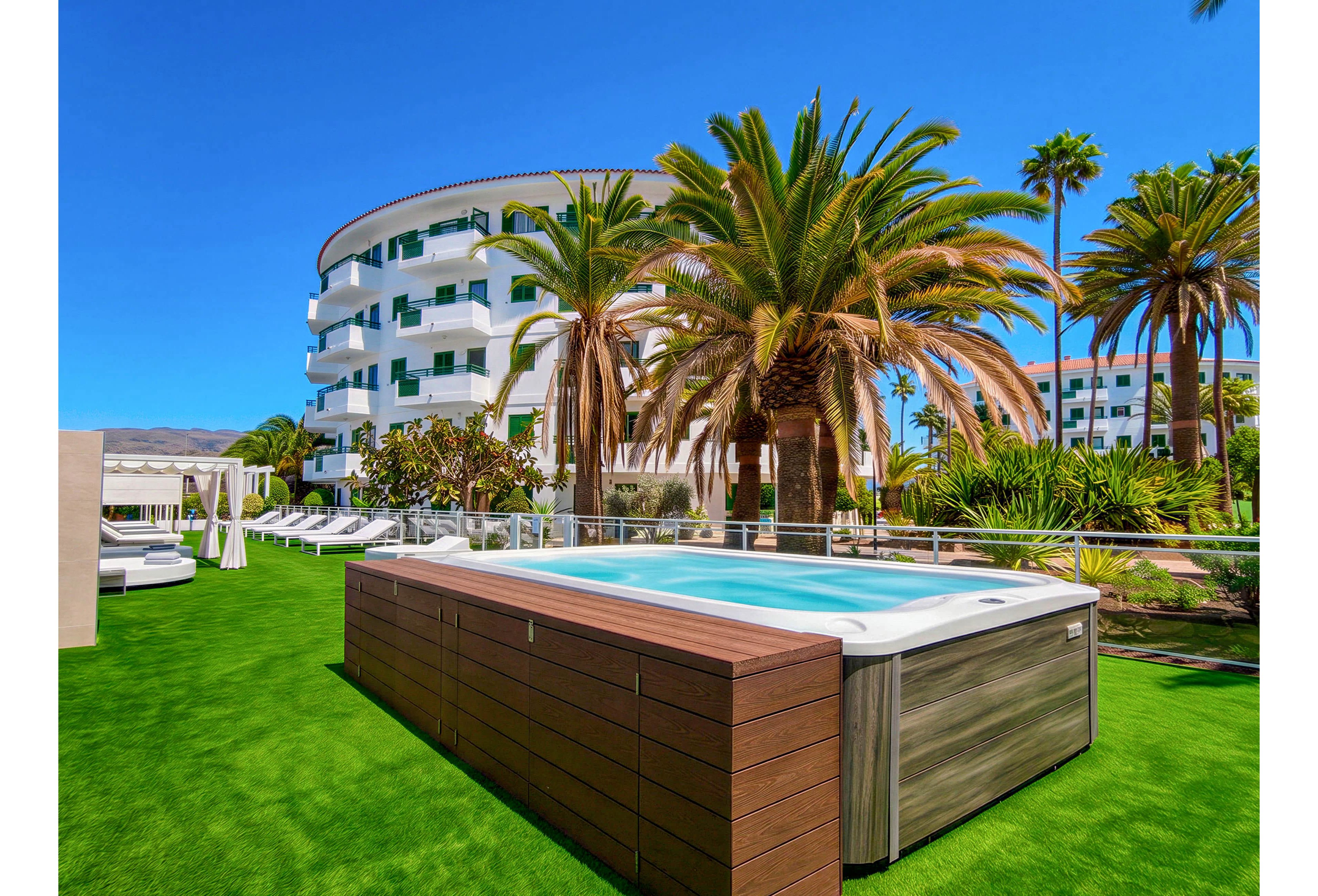 A large rectangular outdoor hot tub sits on bright green artificial grass in a sunny resort courtyard. Behind it, a curved white multi-story hotel building with balconies is visible. Tall palm trees and lush tropical plants surround the area, creating a vacation atmosphere. Lounge chairs and cabanas are arranged nearby under the clear blue sky.
