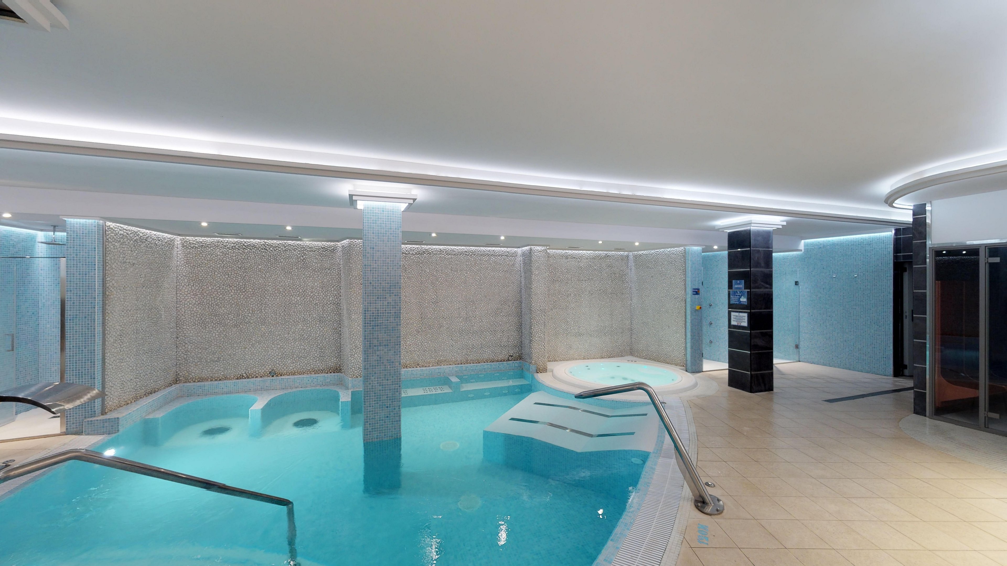 A contemporary indoor spa area features a curved swimming pool with stainless steel handrails. Soft blue lighting and tiled walls create a clean, relaxing atmosphere. The space includes a shallow lounging section and adjacent wellness facilities. The bright ceiling lights and spacious layout emphasize comfort and luxury.