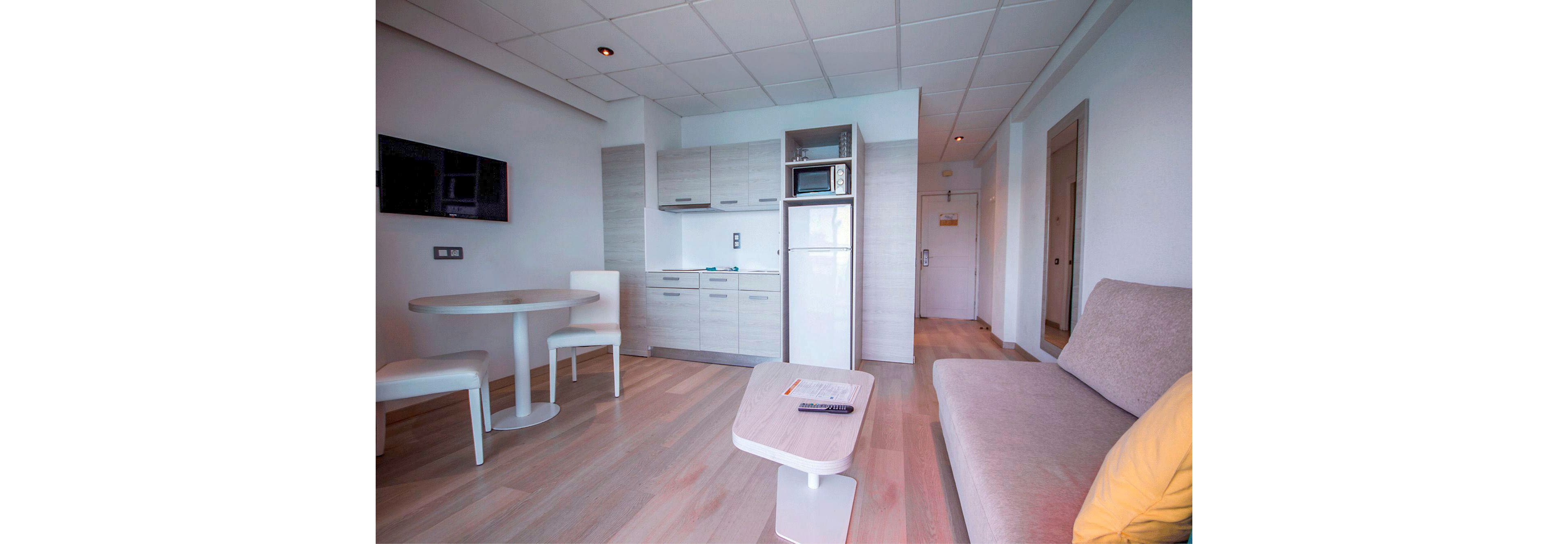Bright modern studio apartment featuring a compact kitchenette with light wood cabinets and a tall white refrigerator. A small round dining table with two chairs sits near the wall-mounted flat-screen TV. In the foreground, a minimalist coffee table holds a remote control and pencil in front of a light sofa with a yellow cushion. The space has pale wooden flooring, white walls, and a clean, contemporary design. A hallway and interior doors are visible in the background, suggesting an entryway and additional rooms.