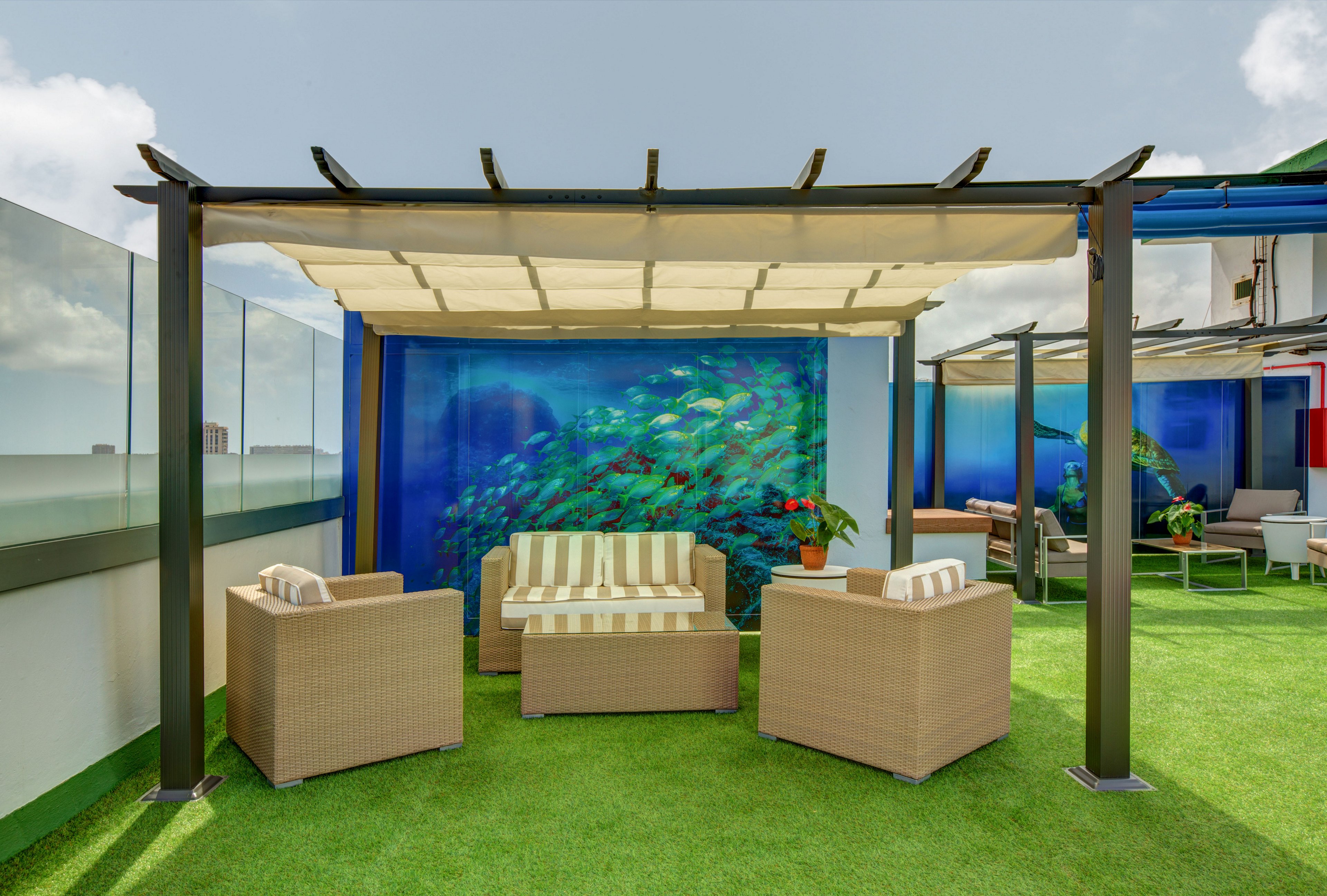An outdoor lounge area features a pergola with a light fabric canopy and wicker-style seating on bright green artificial grass. Two armchairs and a striped cushioned sofa sit beneath the structure, creating a relaxed seating arrangement. Behind the lounge is a vivid ocean-themed mural with colorful fish, adding a playful visual focal point. The setting appears to be on a rooftop or terrace with glass railings and additional seating areas in the background under a partly cloudy sky.