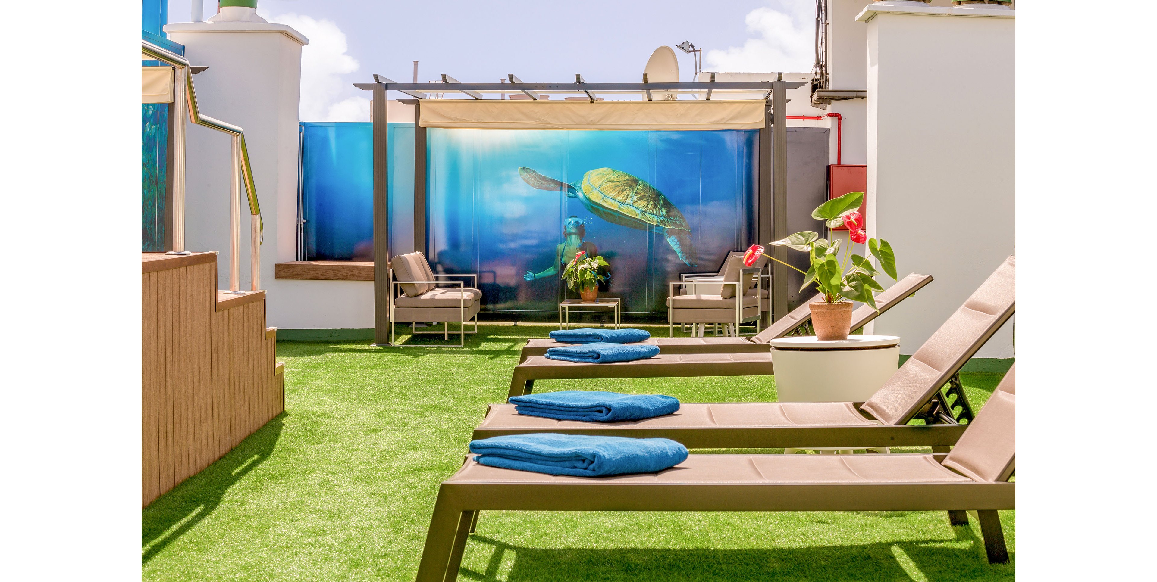 A bright rooftop terrace features several reclining lounge chairs topped with neatly folded blue towels. In the background, a shaded pergola shelters cushioned seating and potted plants. A large underwater-style mural with a swimming sea turtle adds a colorful focal point to the space. The artificial grass flooring and clear sky create a relaxed, resort-like atmosphere ideal for sunbathing or casual conversation.