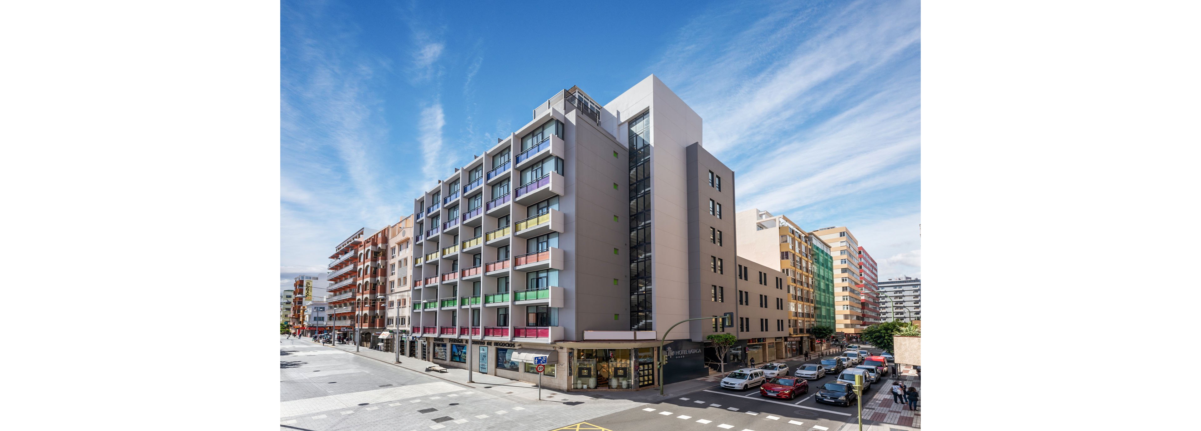 A contemporary multi-story apartment building stands on a busy city corner. Colorful balconies and large windows line the facade, with shops and businesses at street level. Cars are parked along the road and a few pedestrians walk on the wide sidewalk. The scene is captured under a bright blue sky with scattered clouds, emphasizing the vibrant urban environment.