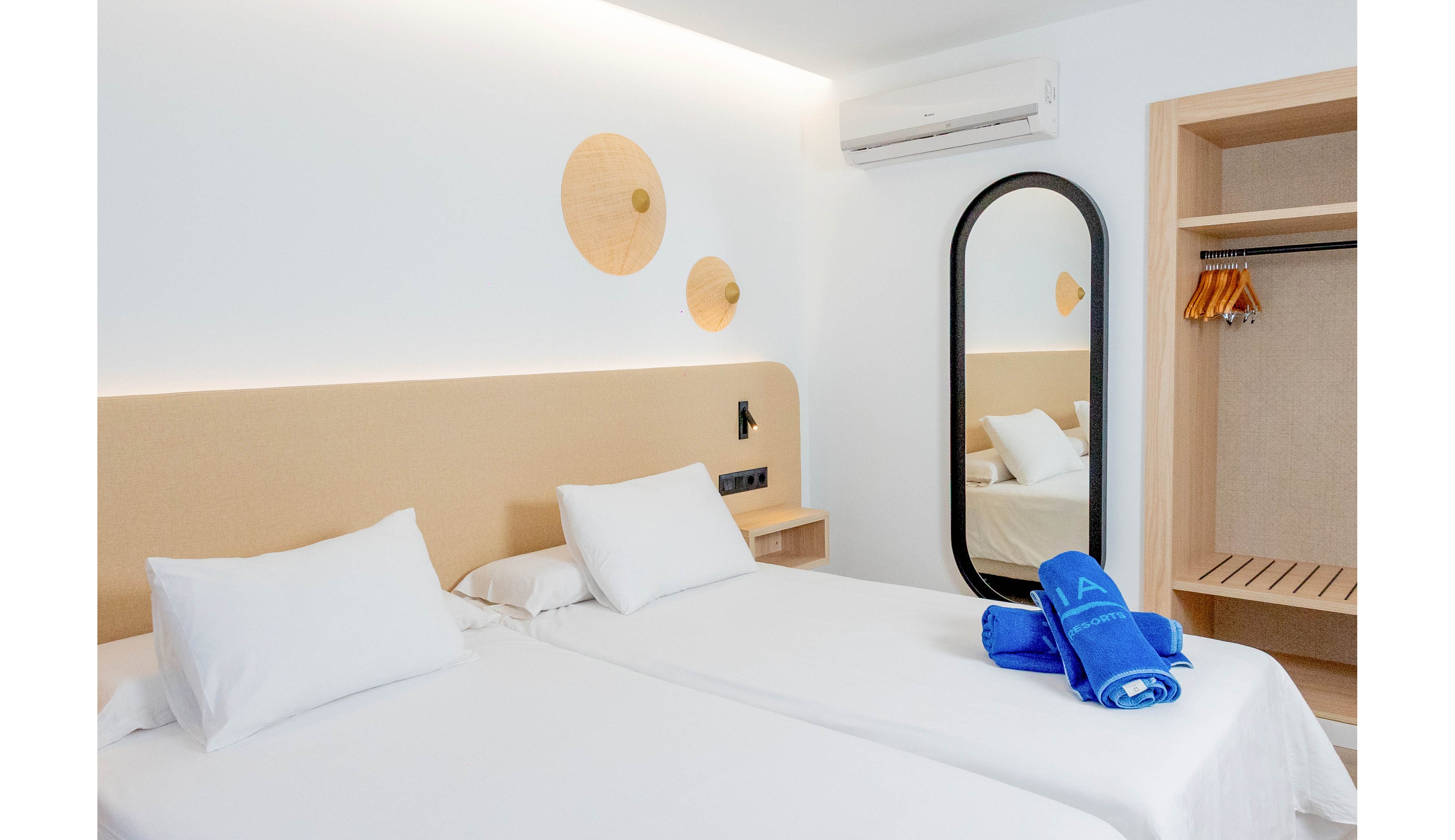 A bright modern hotel room features two neatly made twin beds with white linens and pillows. The space has minimalist decor, including circular wall accents, a black wall lamp, and a tall oval mirror beside an open wardrobe with wooden hangers. Rolled blue towels are placed on one of the beds, adding a pop of color to the neutral palette. An air conditioning unit is mounted above, suggesting a comfortable and climate-controlled environment.