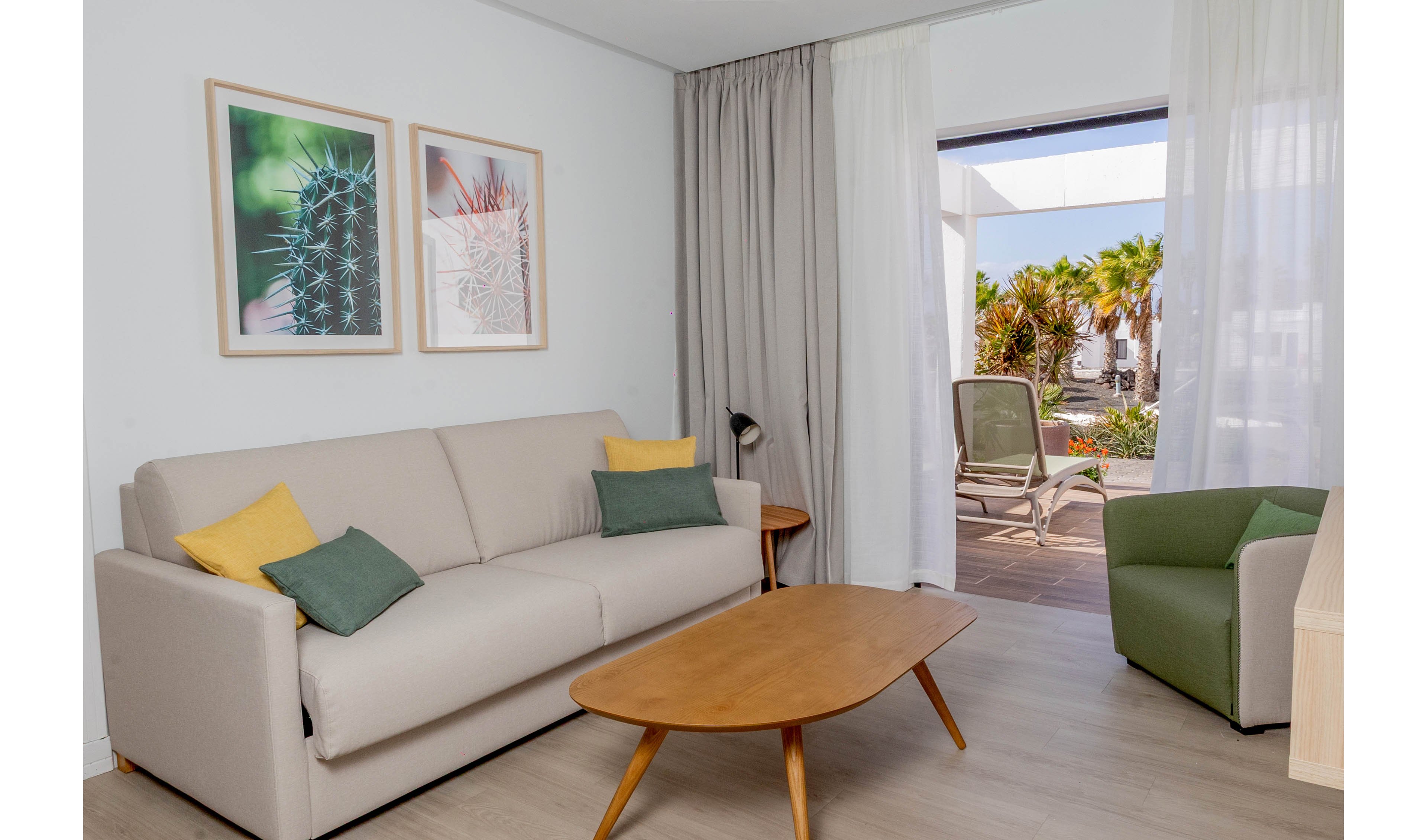 A contemporary living room features a light beige sofa with colorful accent pillows and a wooden coffee table. Two framed cactus prints hang on the wall above the sofa, adding a touch of nature-inspired decor. A green armchair and a small floor lamp complete the cozy seating area. Sheer and opaque curtains frame a large doorway that opens to a sunny terrace with lounge chairs and palm trees outside.