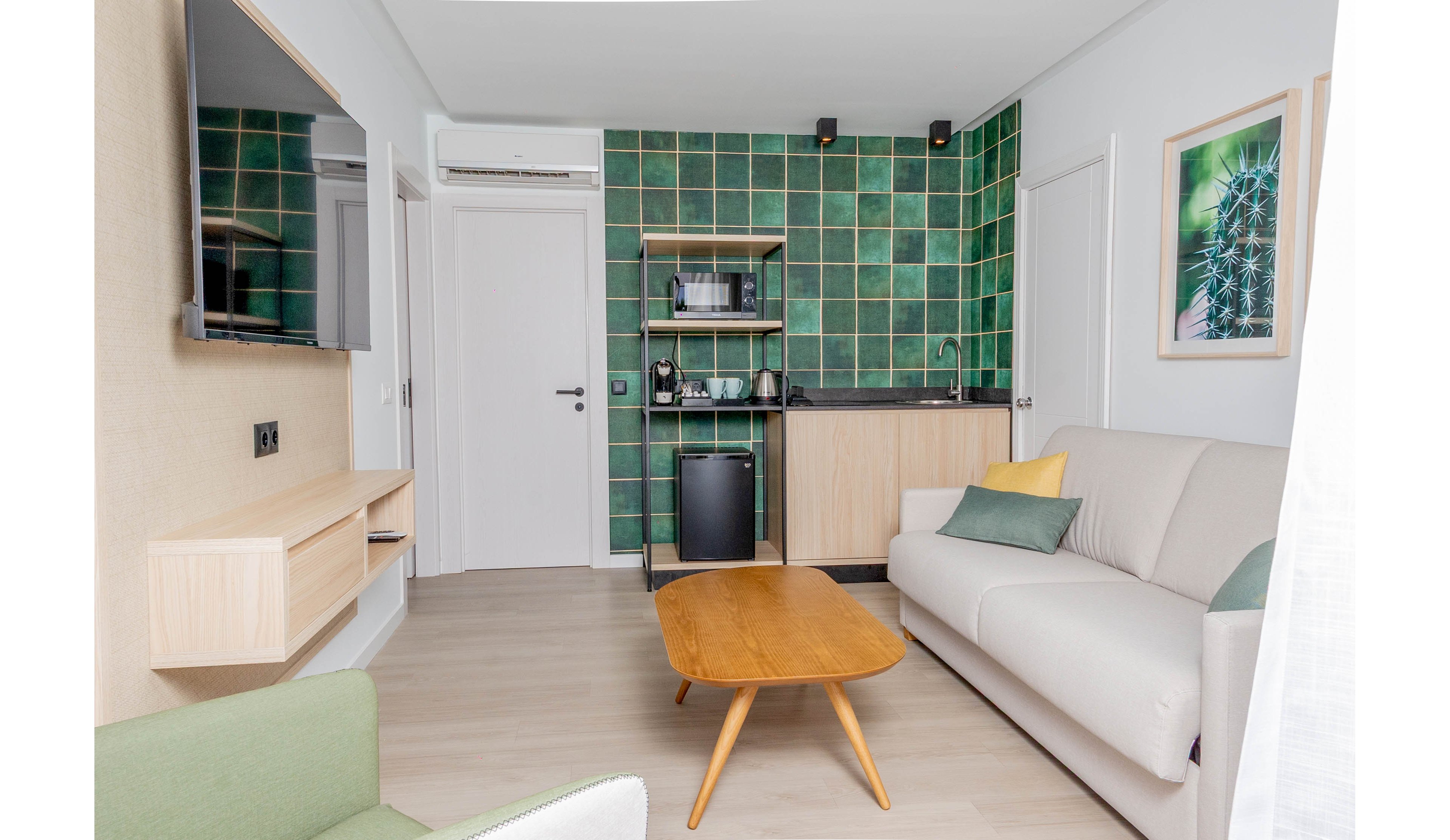 A compact modern living room features a light-colored sofa and wooden coffee table in the foreground. In the background, a small kitchenette with green tiled walls, open shelving, and a mini fridge is neatly arranged. A wall-mounted TV and floating wooden shelves add to the clean, contemporary design. Soft neutral tones with green accents create a calm, minimalist atmosphere.