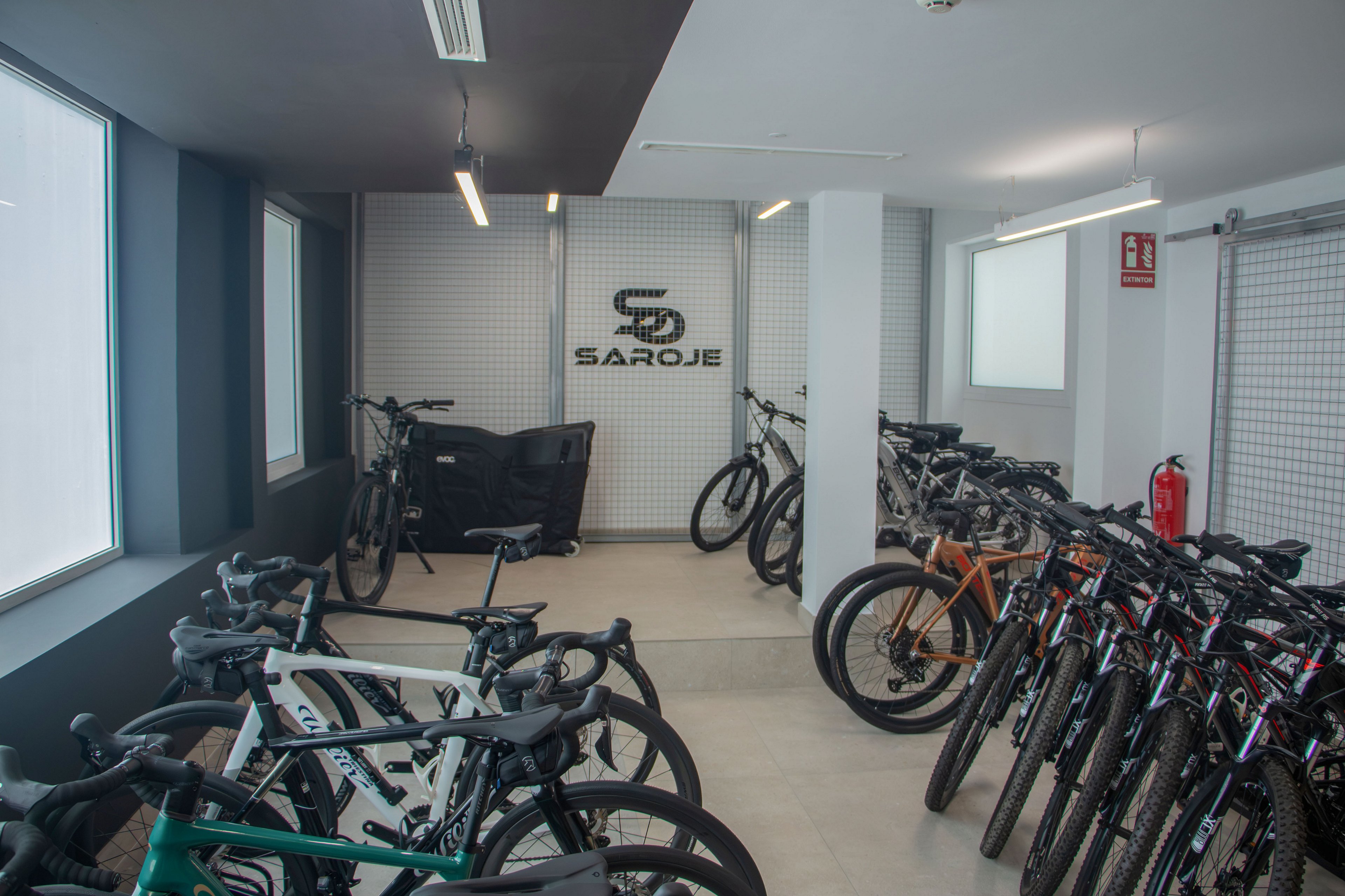 A modern indoor storage room displays multiple bicycles neatly arranged in rows. The space features white walls, large windows, and overhead strip lighting, creating a clean and organized environment. A grid wall at the back shows the text "SAROJE" with a stylized "S" above it, while a fire extinguisher is mounted on the right side. Several mountain and road bikes of different colors and styles are visible throughout the room.