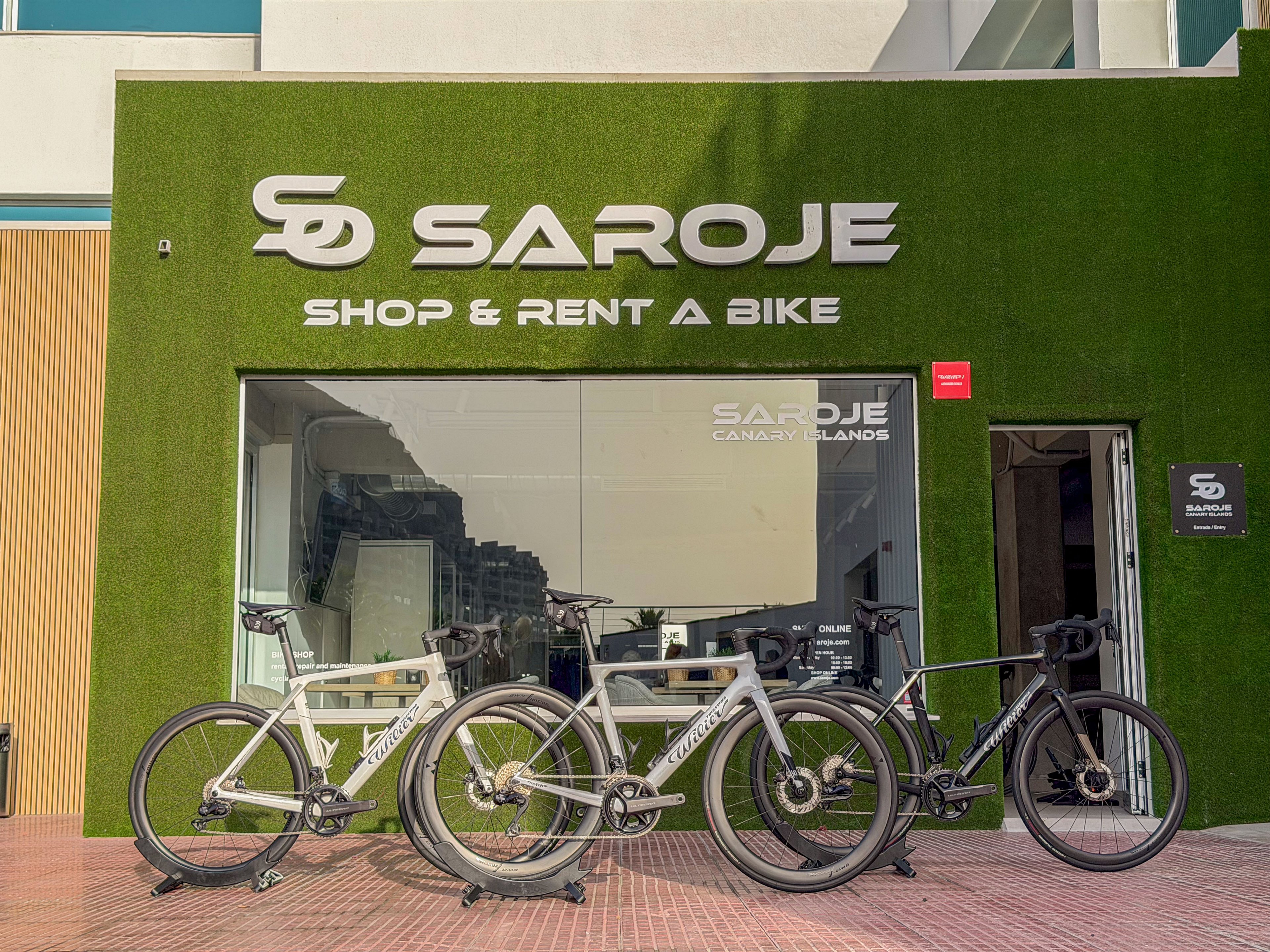 Three high-performance road bicycles are displayed outside the Sarojé shop, which offers bike rental services. The storefront features a green grass-like facade with large metallic lettering reading 'SAROJE' and 'SHOP & RENT A BIKE'. A glass window reveals the interior of the store and reflections of nearby buildings. The scene is captured in daylight on a paved sidewalk, emphasizing an urban cycling environment.