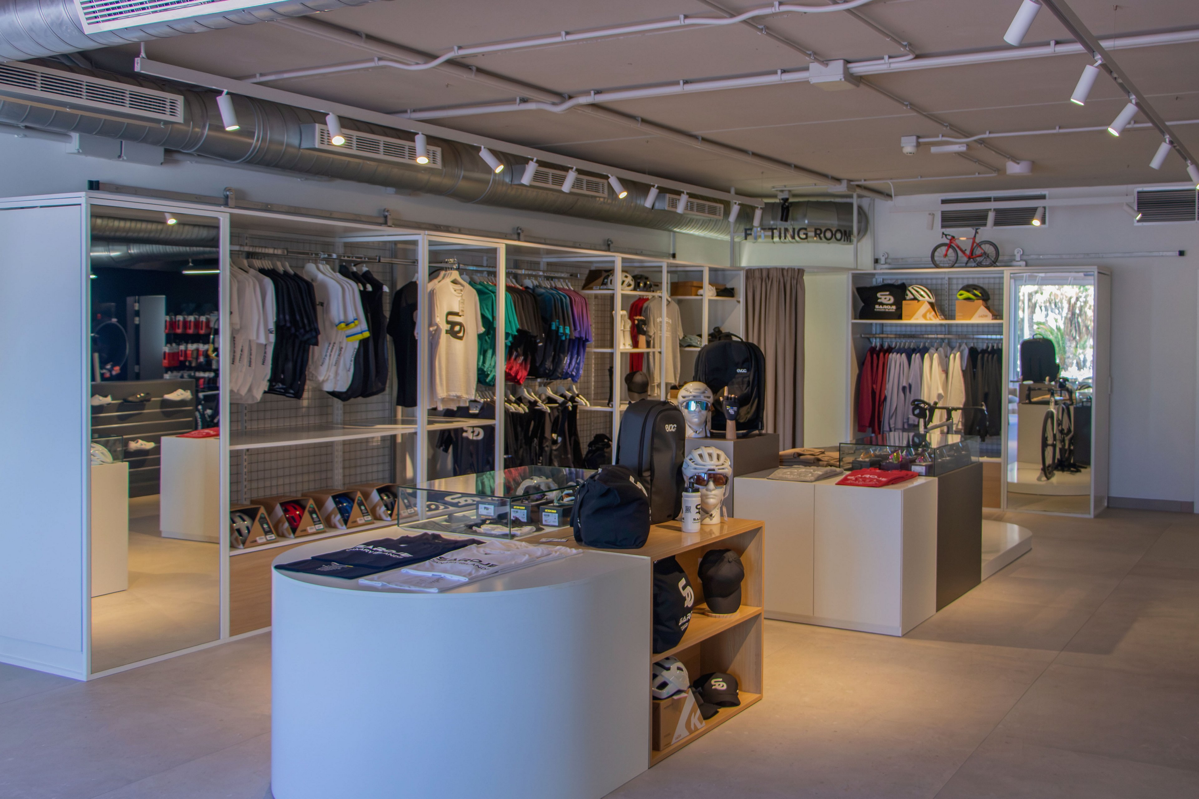 Contemporary retail interior featuring sports apparel and accessories neatly displayed on racks and shelves. T-shirts, jackets, and athletic clothing are organized along the walls, with bags and smaller items arranged on central white display units. The store has a clean, minimalist design with neutral flooring and exposed ceiling fixtures. Bright, even lighting and spacious aisles create an inviting shopping environment focused on active lifestyle products.