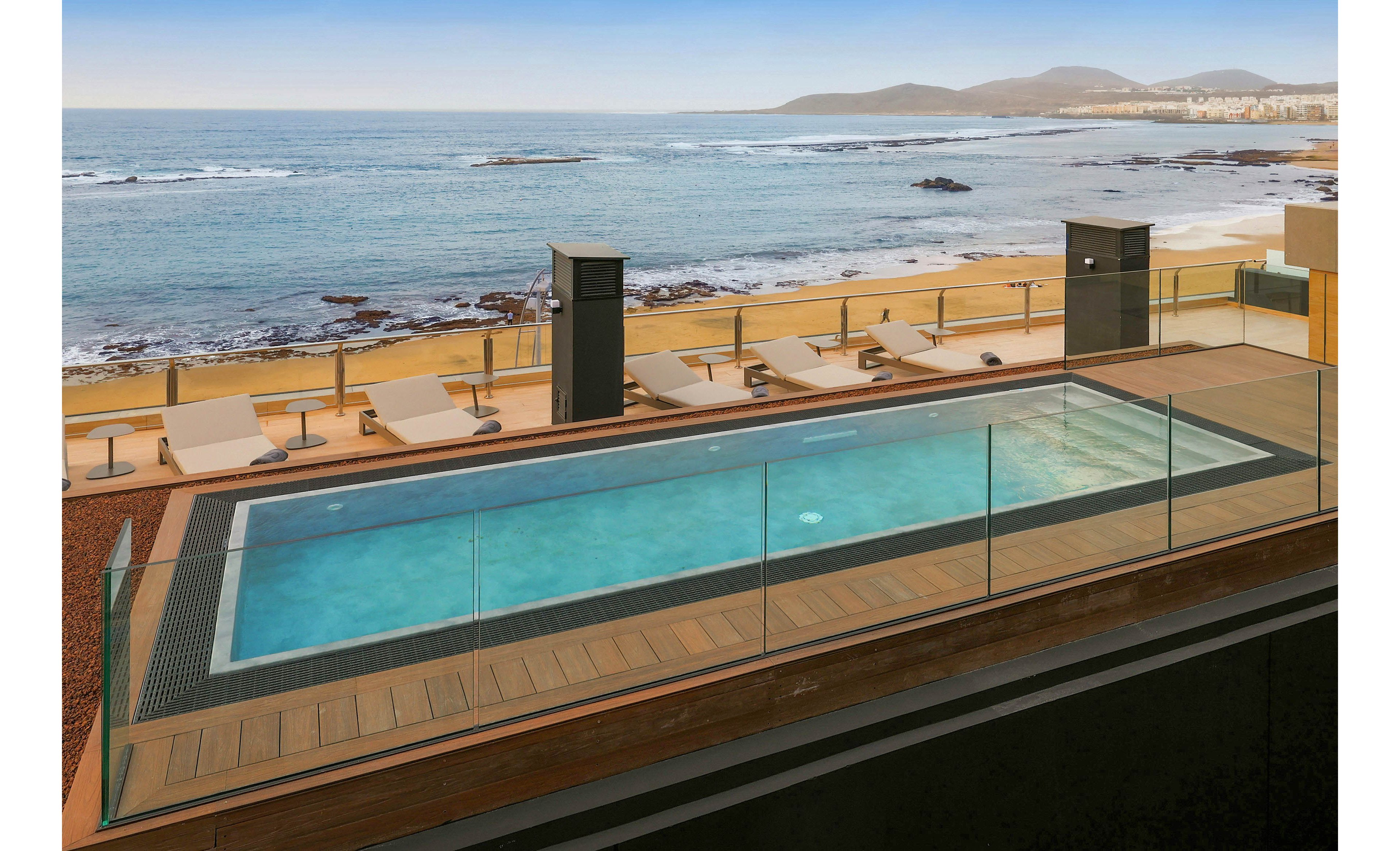 A rectangular rooftop swimming pool is surrounded by a wooden deck and glass safety railing. Several beige lounge chairs line the deck, facing the ocean and sandy beach below. Calm blue water stretches to the horizon with gentle waves and distant hills under a clear sky. The scene suggests a tranquil, contemporary seaside resort ideal for relaxation and sunbathing.