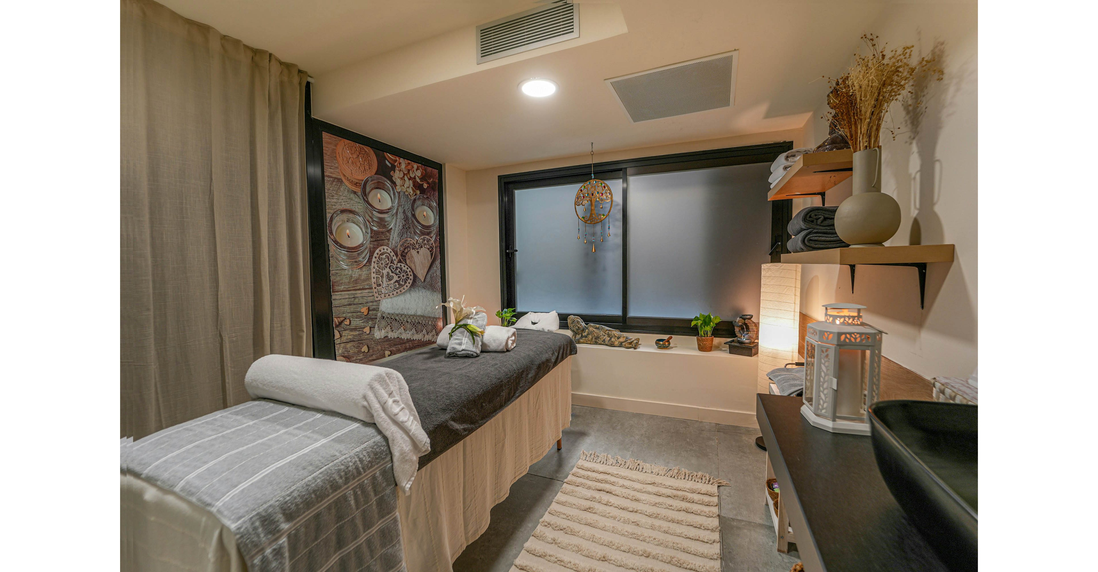 A neatly prepared massage table is the focal point of a cozy spa treatment room. Soft lighting, neutral tones, and decorative elements such as a wall mural and hanging ornament create a relaxing atmosphere. Towels, candles, and small plants are arranged around the space, enhancing the sense of calm and comfort. The room appears ready for a wellness treatment or therapeutic massage.