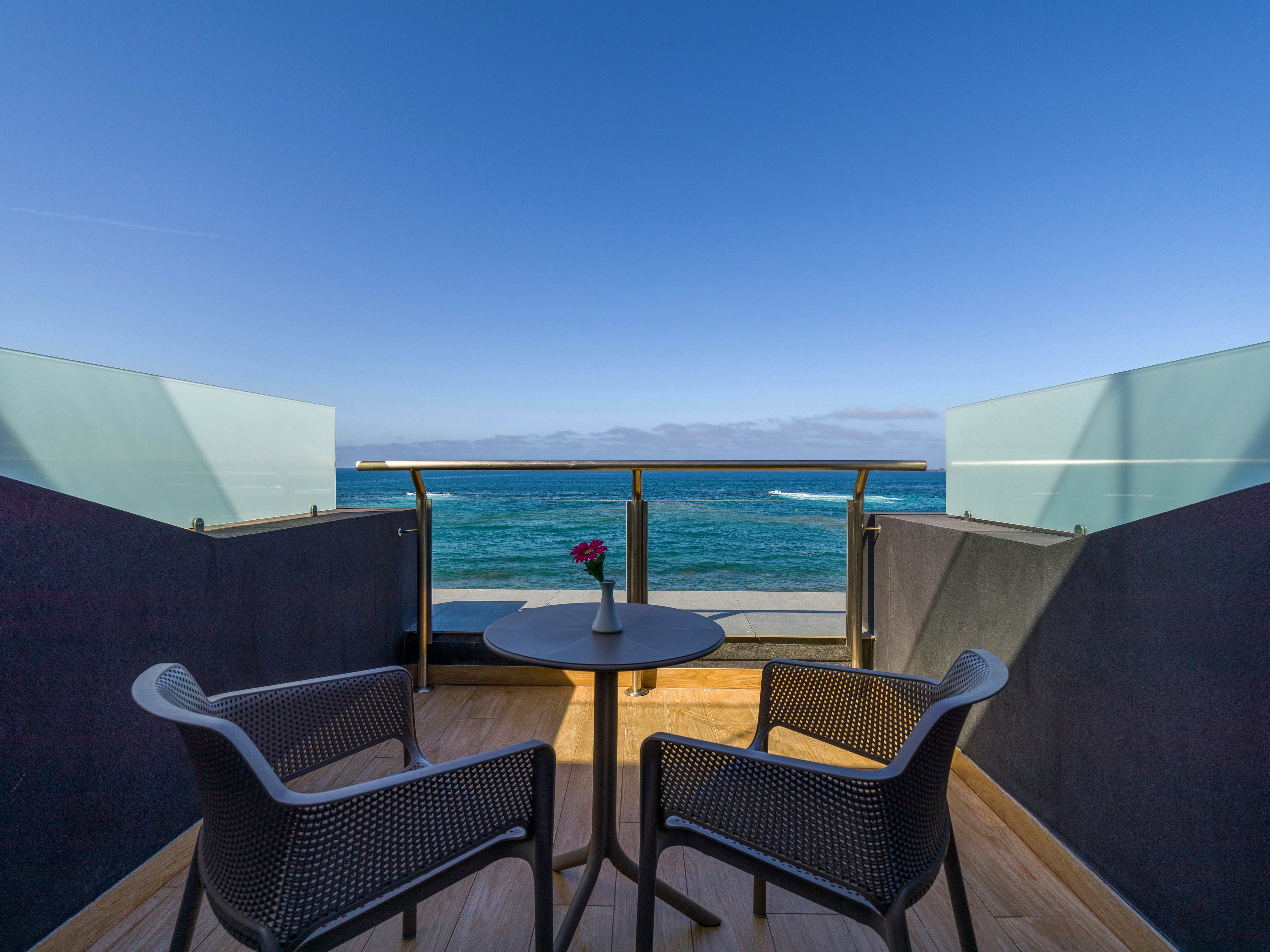 A small balcony with two modern chairs and a round table faces a tranquil ocean view. A single flower in a vase sits on the table, adding a touch of color to the minimalist setting. Glass panels and railings frame the balcony, opening out to clear blue sky and gentle waves. The scene suggests a peaceful coastal retreat ideal for relaxation or conversation.