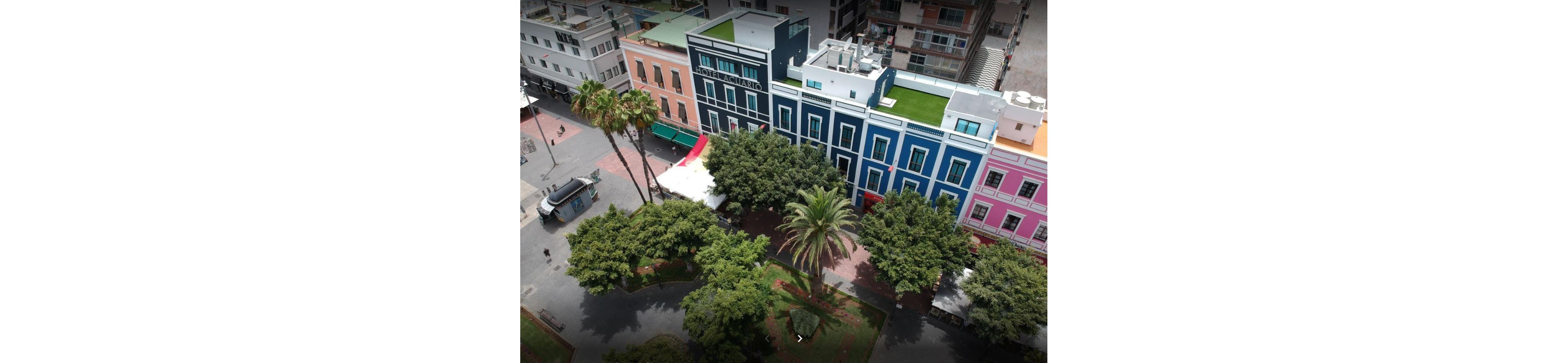 Aerial view of a vibrant city block with colorful multi-story buildings facing a tree-filled urban park. The facades include blue, pink, and pastel tones, with one building featuring a rooftop terrace and green roof. Tall palm trees and dense foliage dominate the park area, with paved walkways and a few people visible below. Surrounding structures and streets frame the scene, highlighting a lively downtown environment.