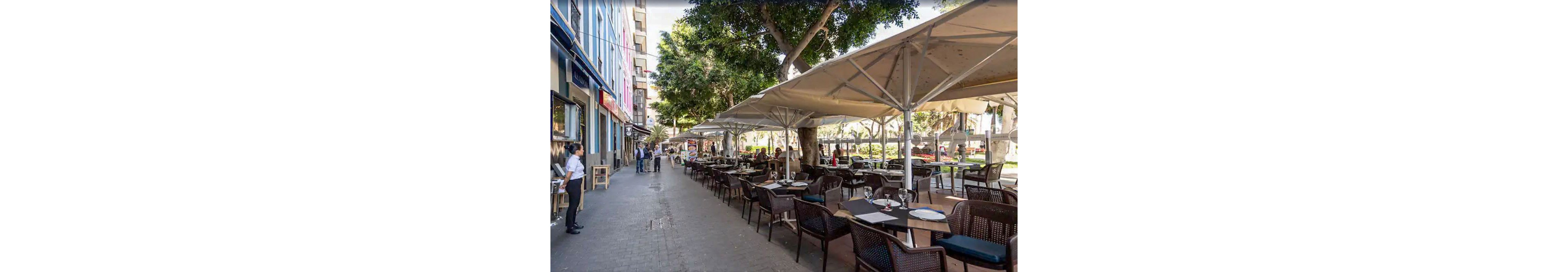 An outdoor café terrace lines a city street with rows of dark wicker chairs and tables set with plates and glasses. Large white umbrellas provide shade beneath tall trees, creating a relaxed dining atmosphere. A server stands near the entrance while a few pedestrians walk along the sidewalk. Buildings with colorful facades and ground-floor businesses frame the urban scene.