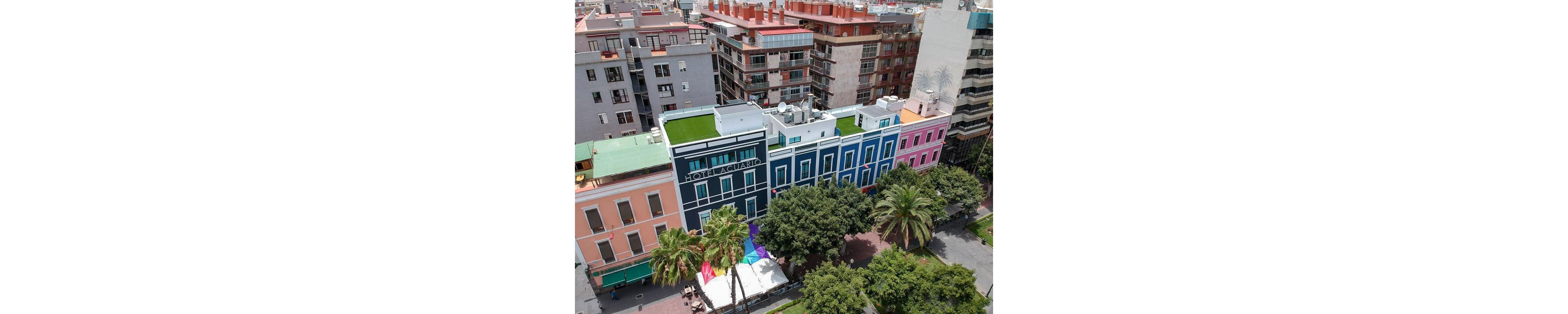 Aerial view of a vibrant city street featuring the Hotel Aguacate with a bold blue facade and rooftop terrace. The hotel is flanked by pastel-colored neighboring buildings and surrounded by lush green trees and a tall palm. A large rainbow-colored fabric or flag is displayed near the entrance, adding bright accents to the scene. In the background, multi-story residential buildings with balconies and rooftop structures complete the urban landscape.